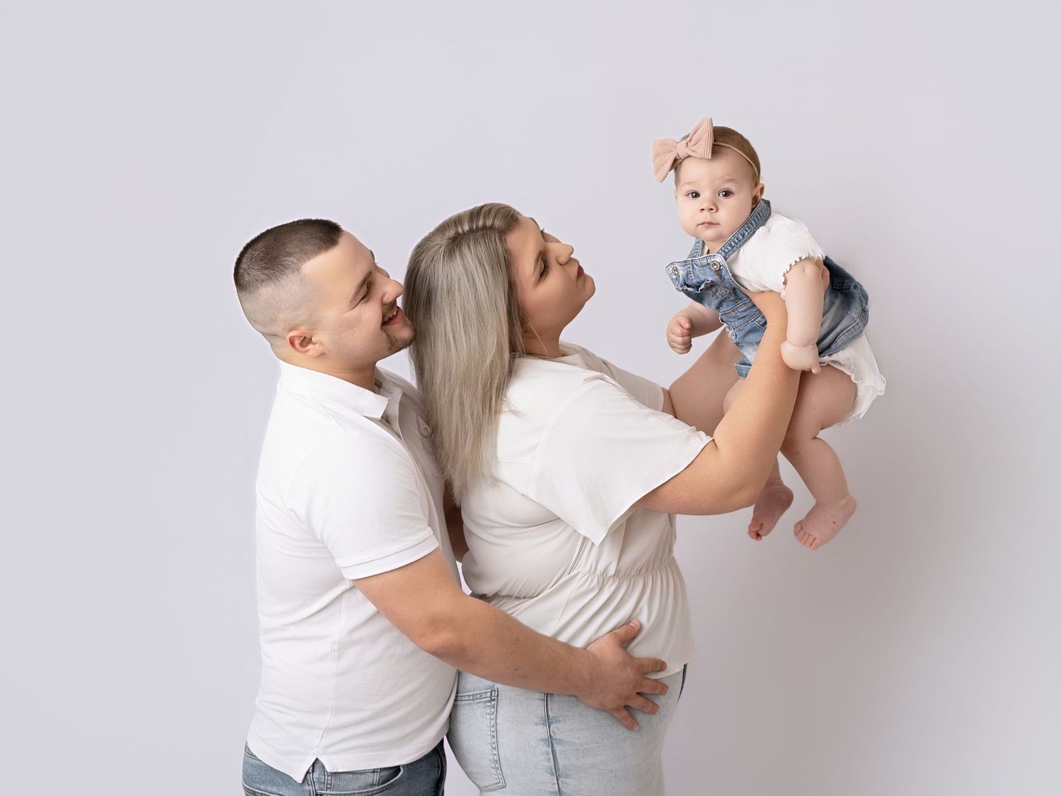 A happy family with a man and woman in white shirts holding their baby girl in a bright studio photo.