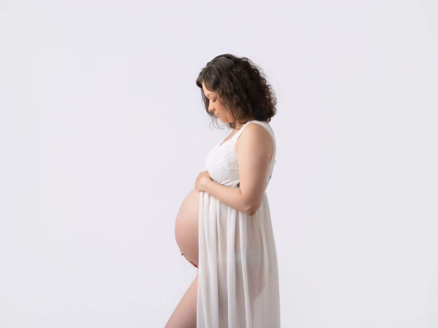 Pregnant woman in white sheer dress cradling her baby bump against a clean white background.