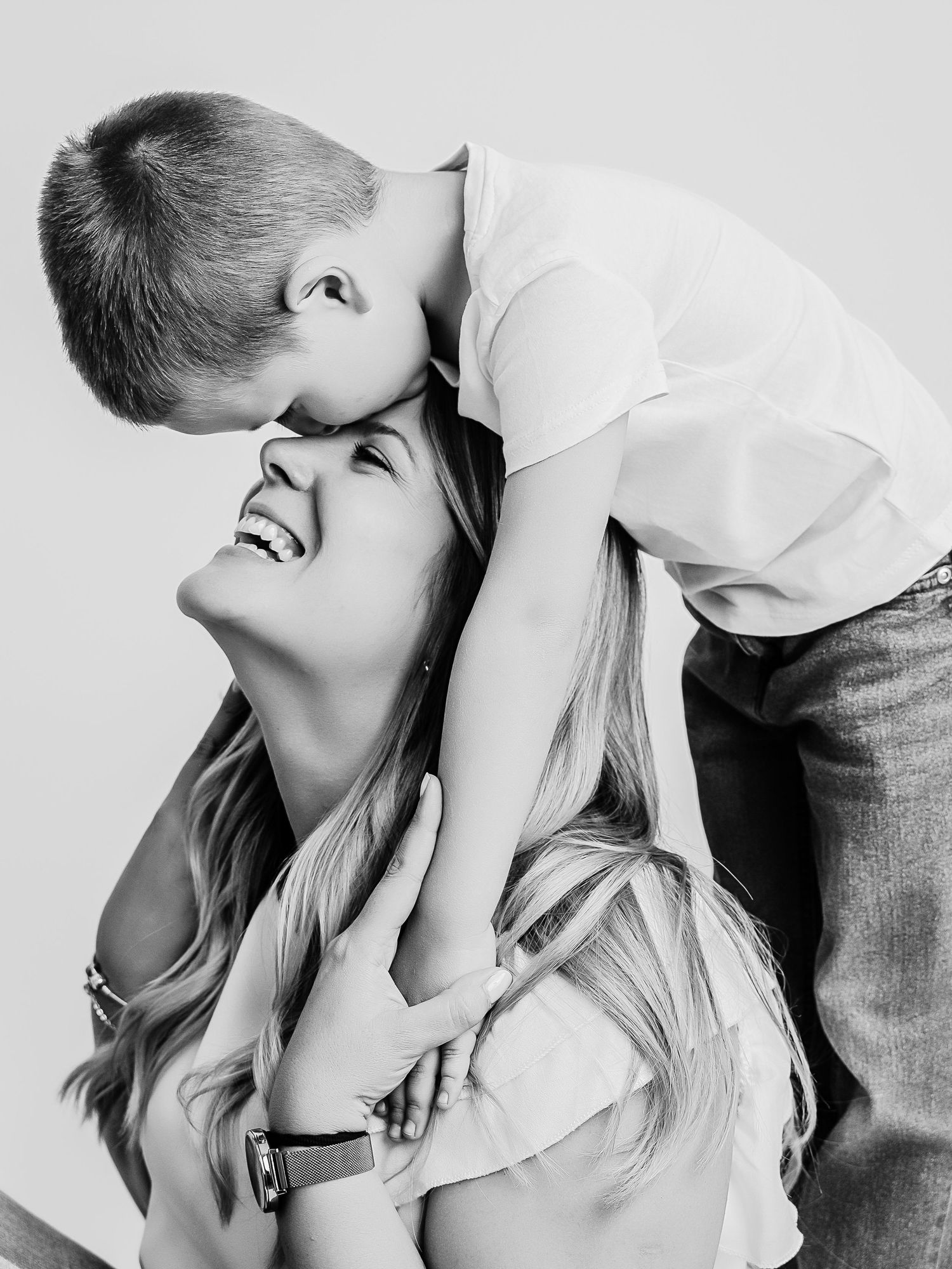 Black and white photo of a laughing mother as her young son leans over and kisses the top of her head.