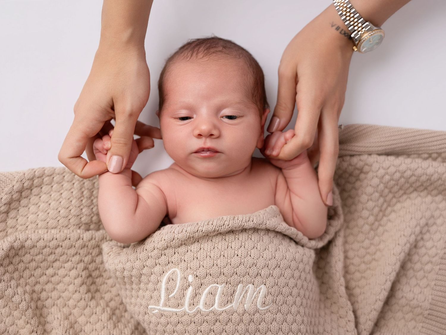 Newborn baby Liam wrapped in a beige knit blanket with embroidered name, cradled by gentle hands.