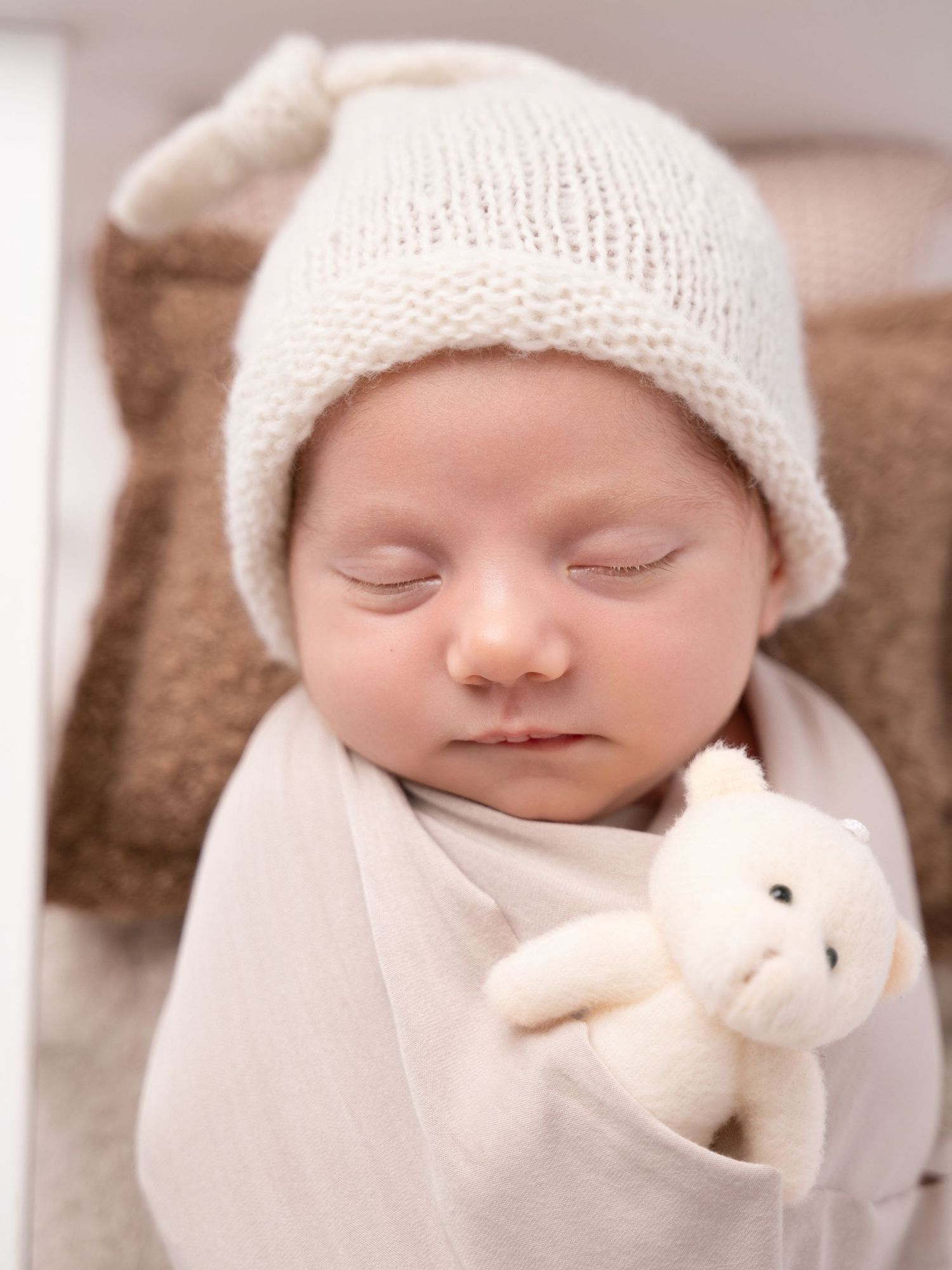 Peaceful newborn baby sleeping wrapped in white swaddle, wearing knit hat, holding tiny white teddy bear stuffed animal.