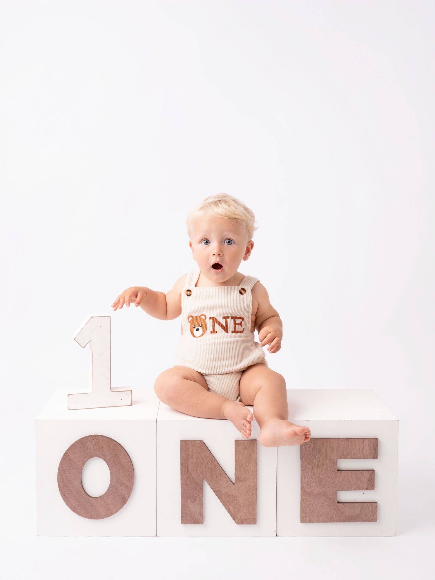 A surprised baby sits on white blocks spelling ONE, wearing a bear onesie, celebrating their first birthday.