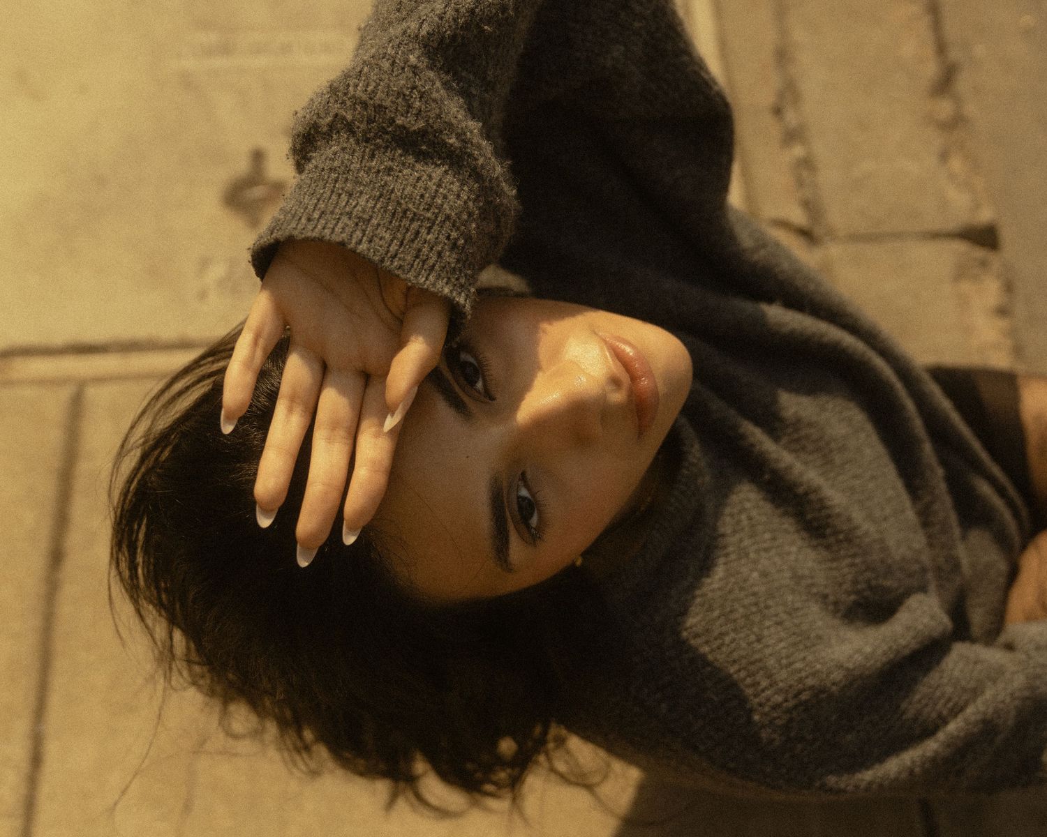 Young woman in gray oversized sweater lying on floor, looking up at camera with hand near face.