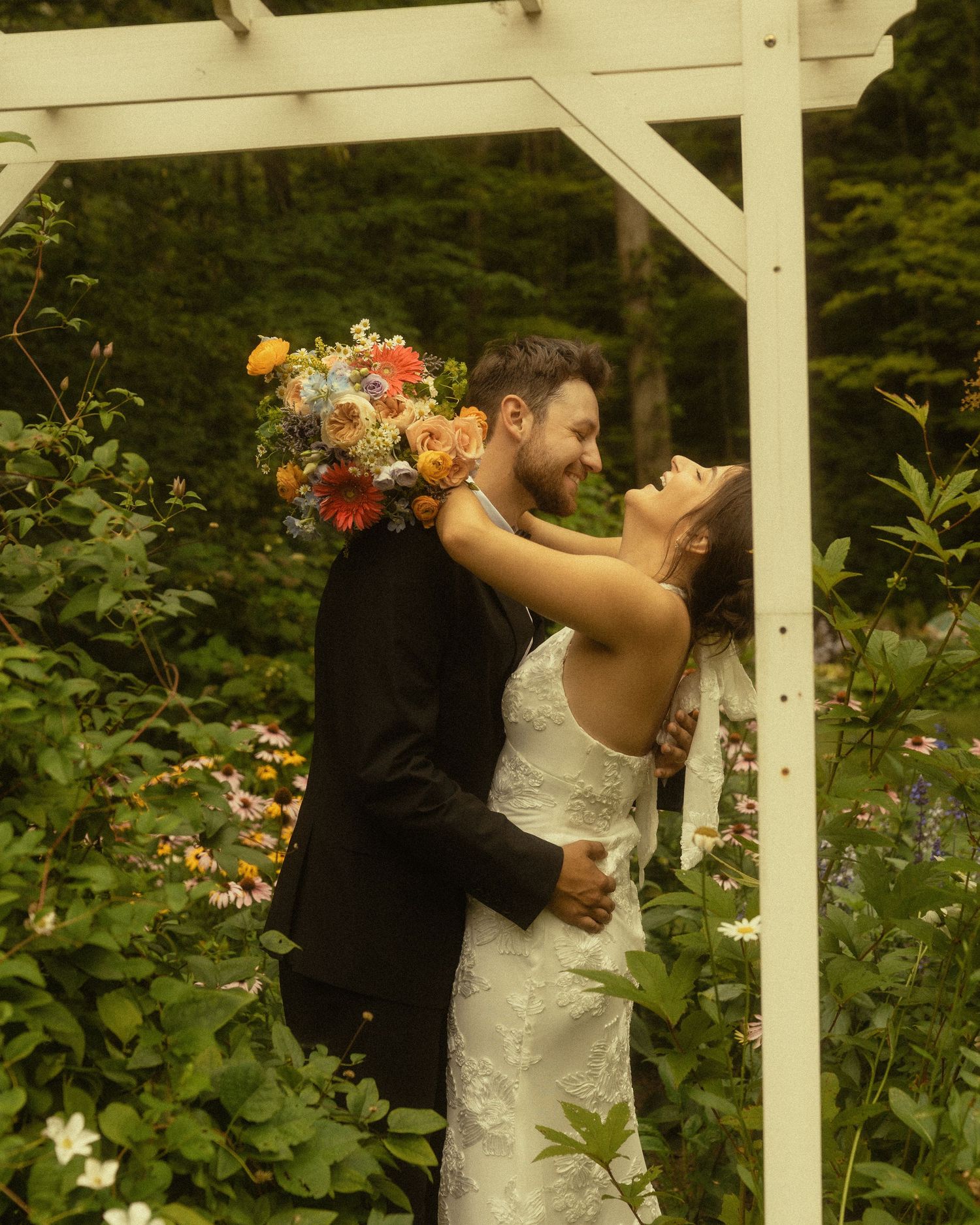 A bride and groom embrace under a white arbor surrounded by lush greenery, the bride holding a vibrant wildflower bouquet.