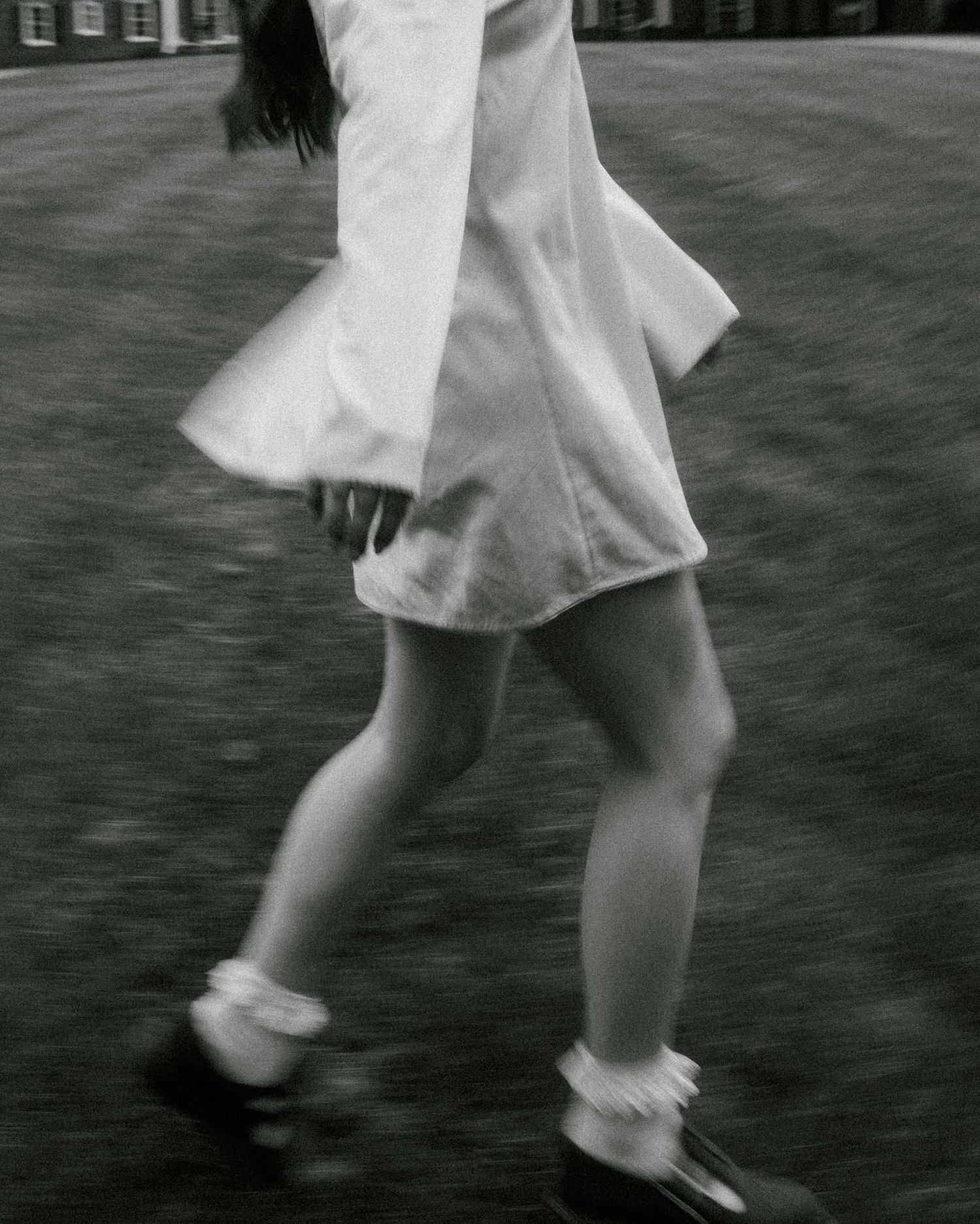 Black and white photo of a girl in white dress and mary janes spinning on grass near a brick building.