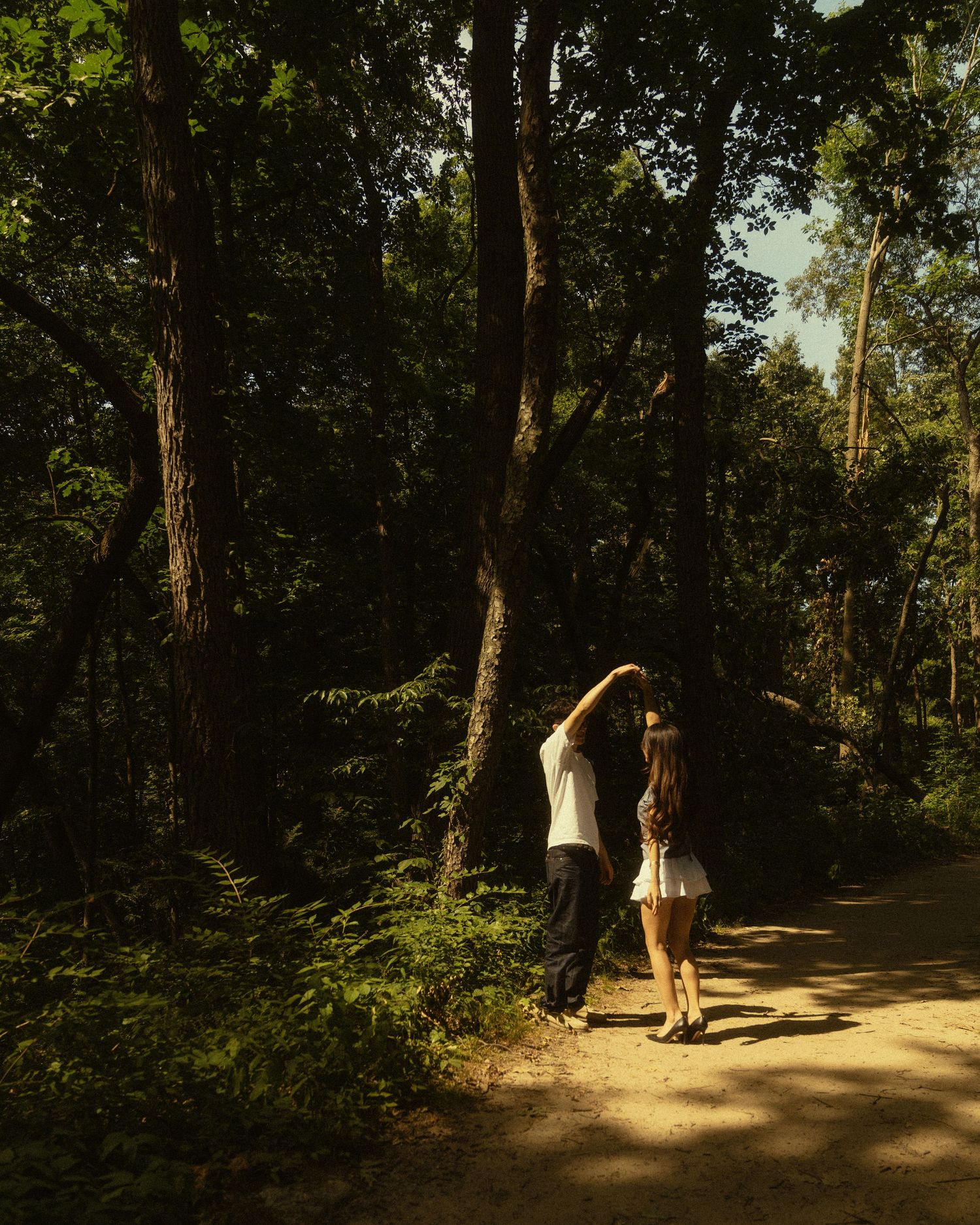 A couple holds hands on a sunlit forest path surrounded by tall trees and lush green foliage.