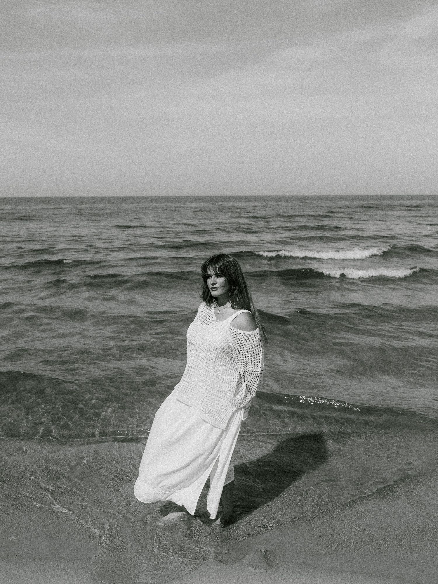 Black and white photo of a woman in a white dress standing in ocean waves on a beach.