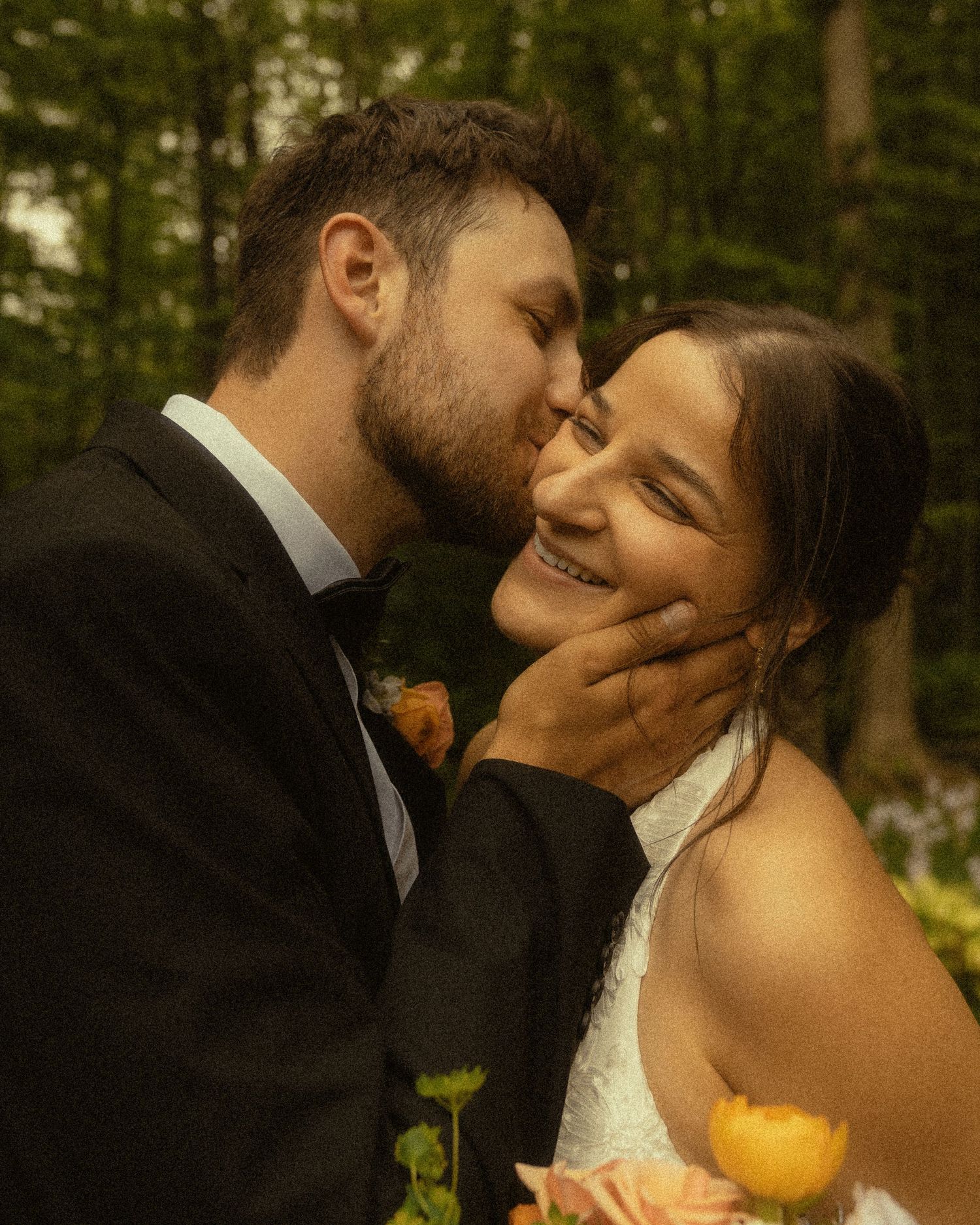 Groom in black tuxedo kissing bride on cheek as she smiles, holding yellow flowers in a forest setting.