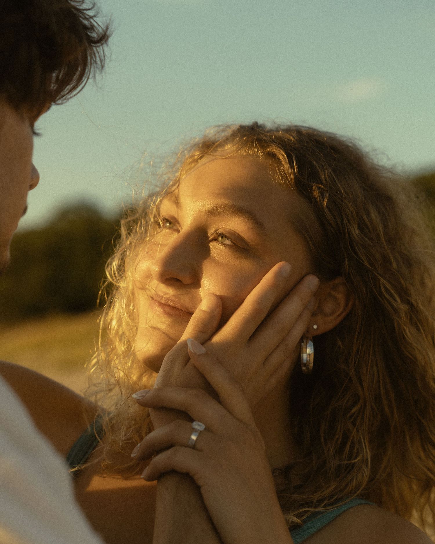 A young woman with curly hair gazes up in golden sunlight, her face cradled in hands wearing a silver ring.