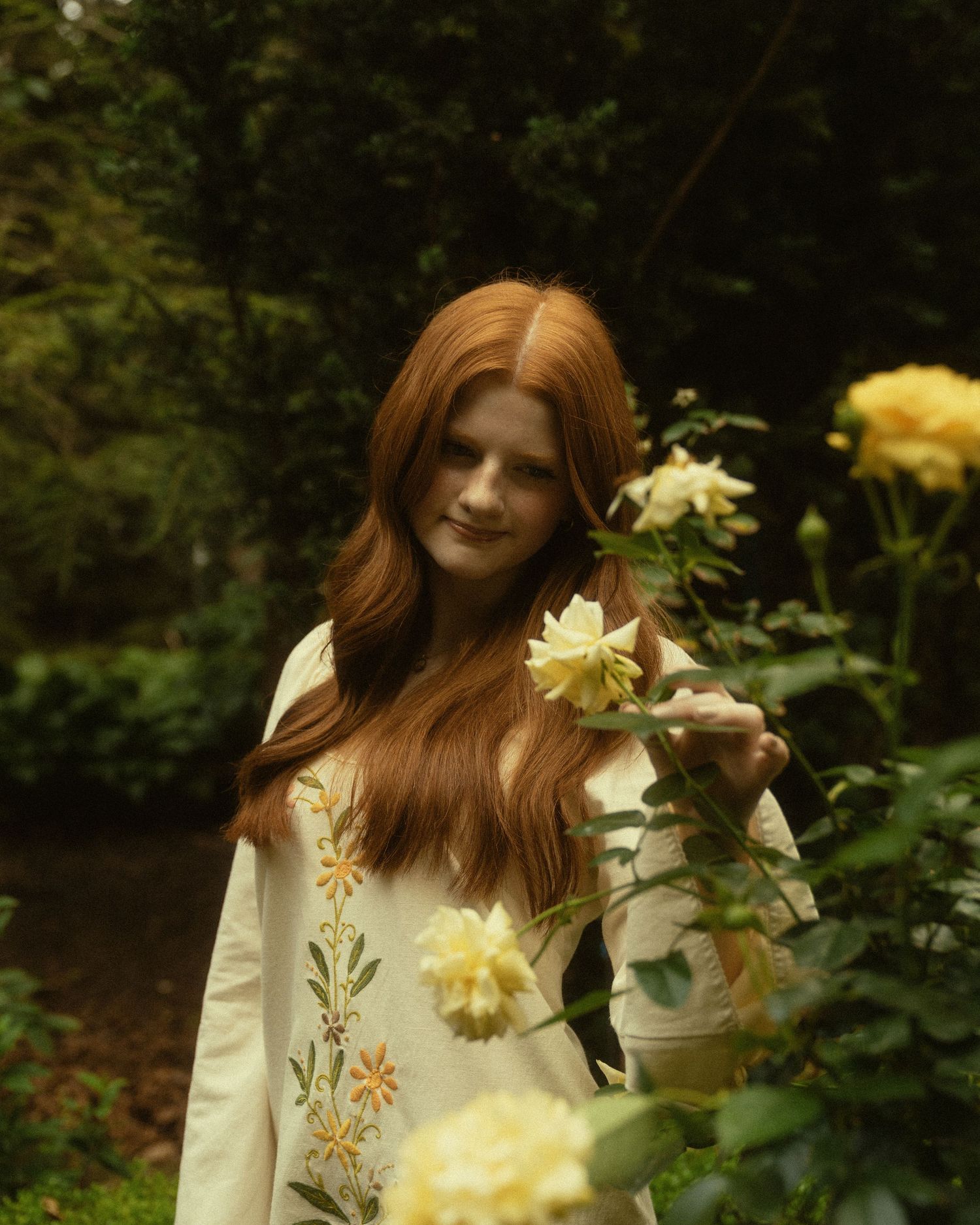 A young woman with long red hair in a floral white dress stands among yellow roses in a lush green garden.