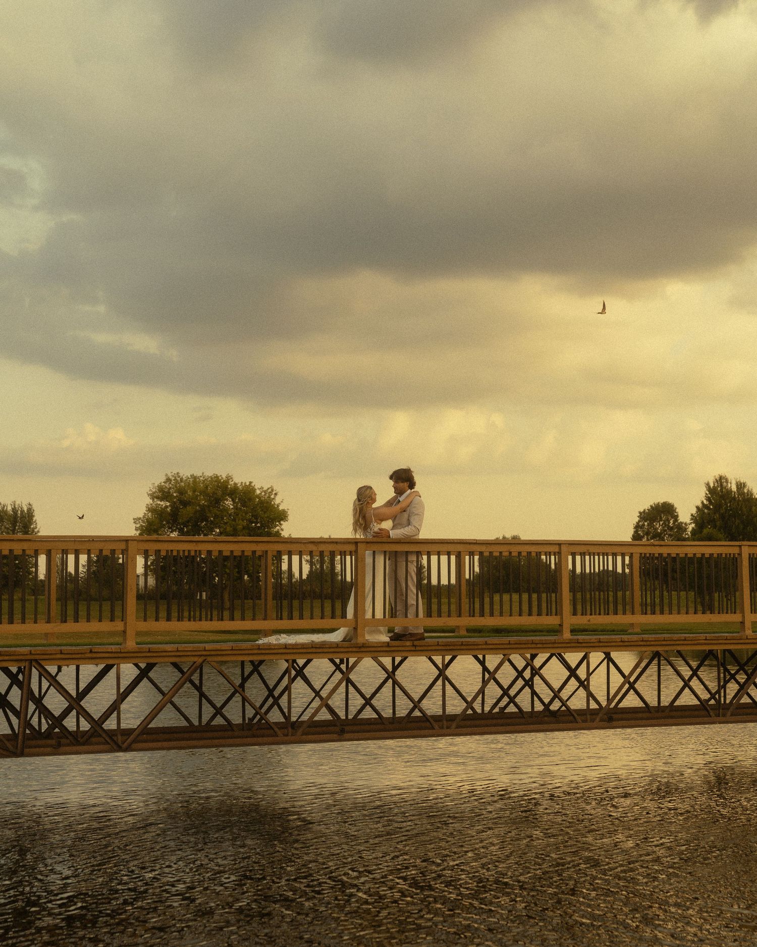 Couple embracing on a wooden bridge over water at golden sunset with dramatic cloudy sky.
