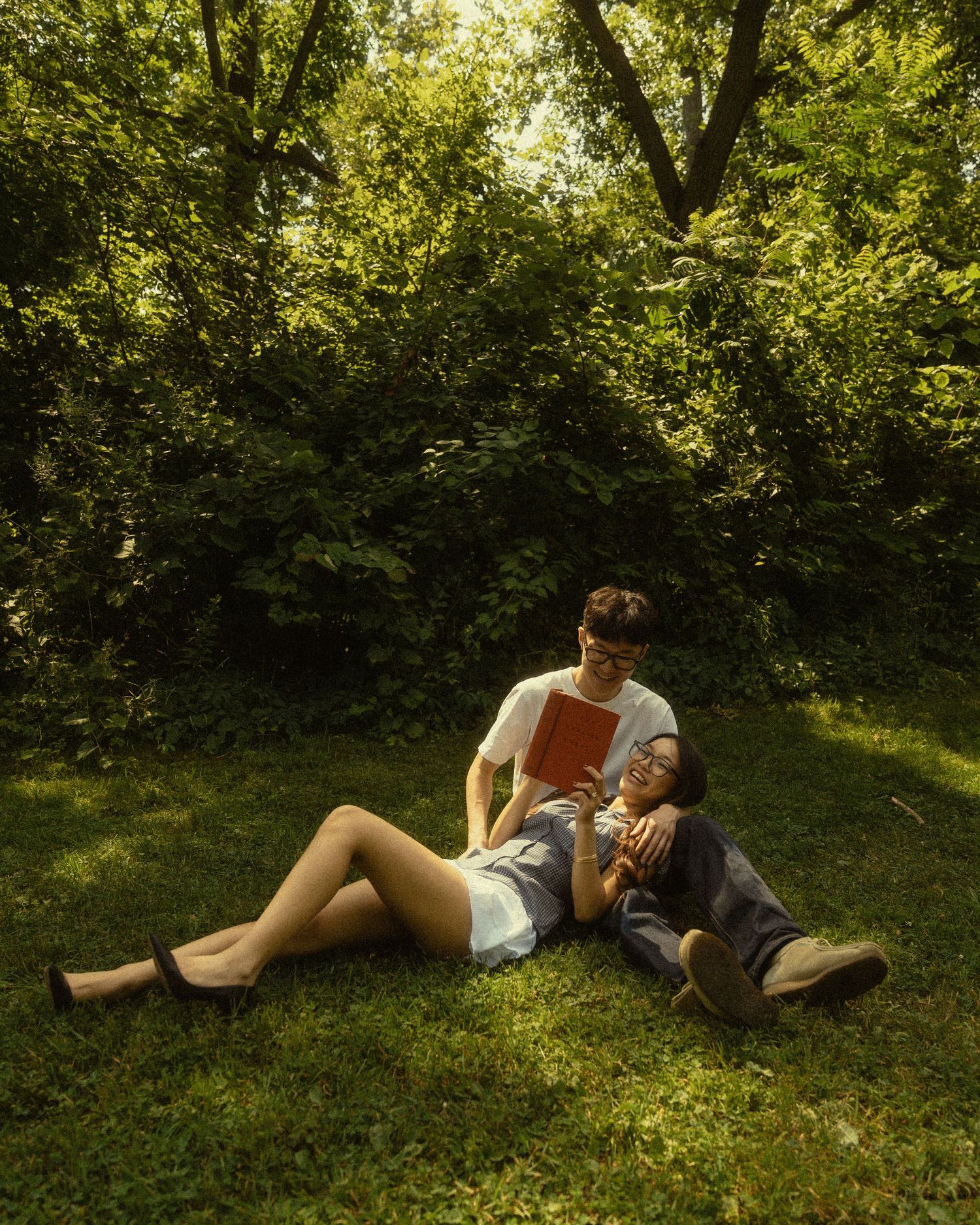 Couple relaxing on green grass in a sunlit park, man holding a book while woman leans against him.