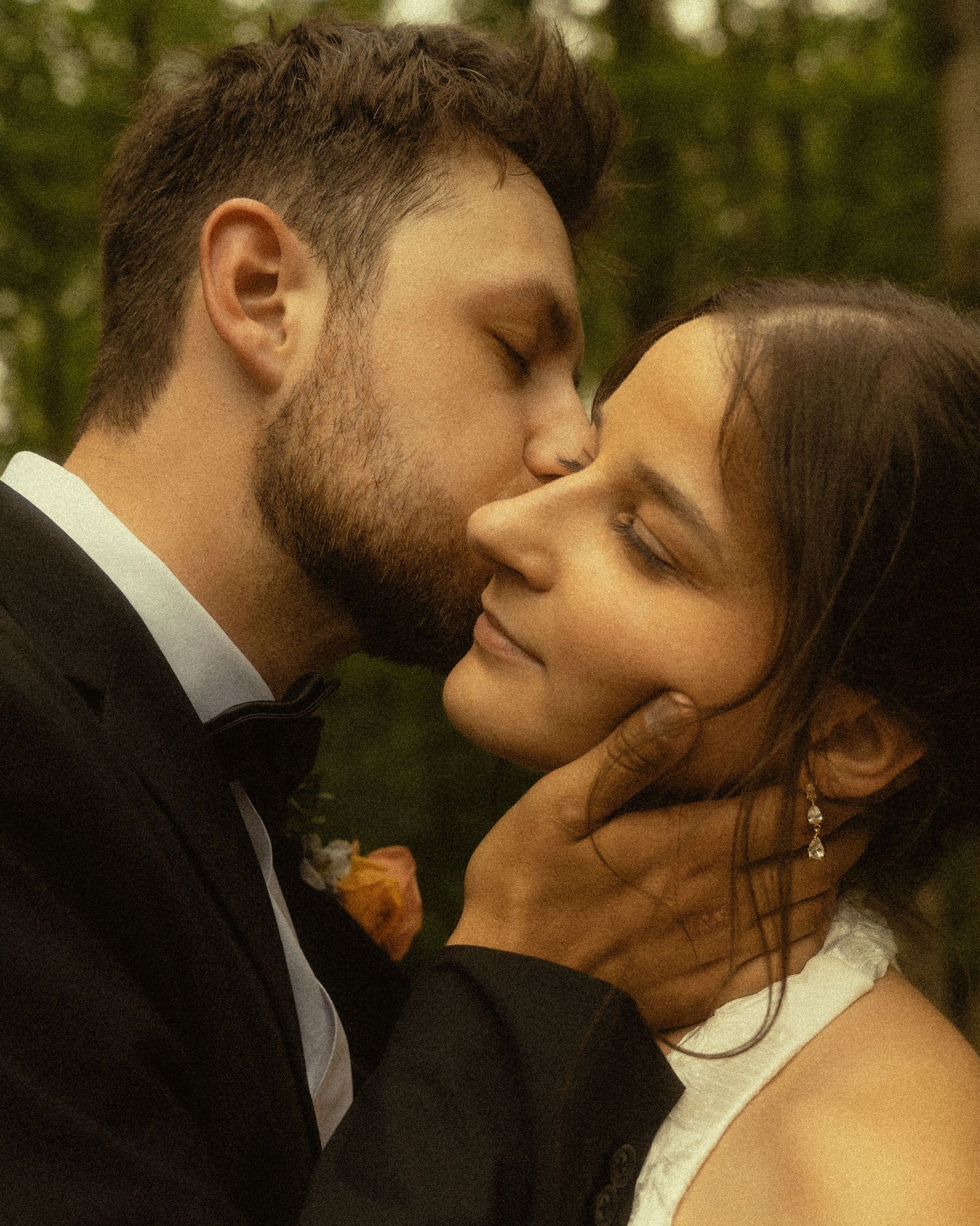 A groom in a black tuxedo tenderly kisses his bride on the cheek during their outdoor wedding ceremony.