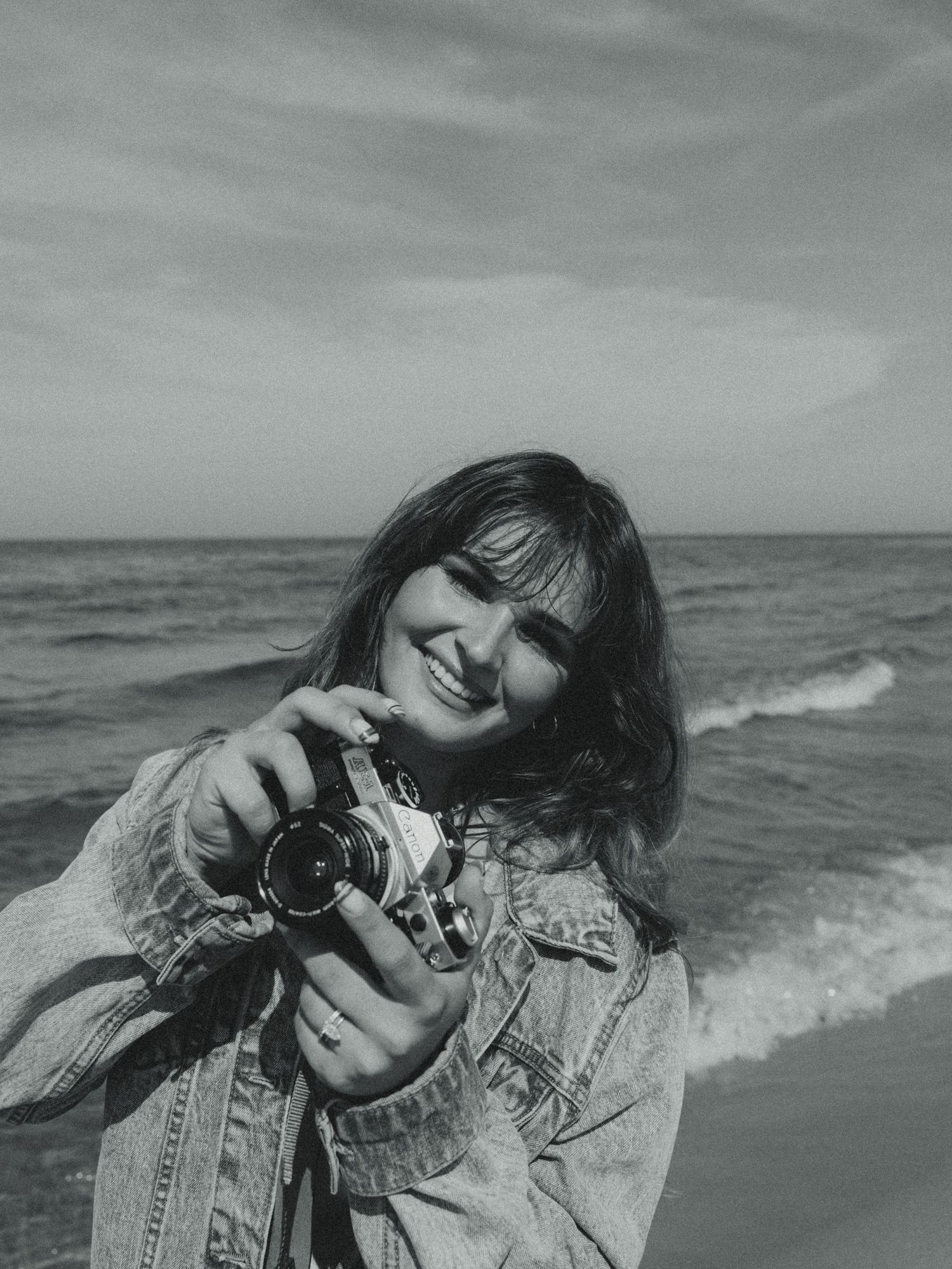 Black and white photo of a smiling young woman holding a vintage camera at the beach with ocean waves behind her.