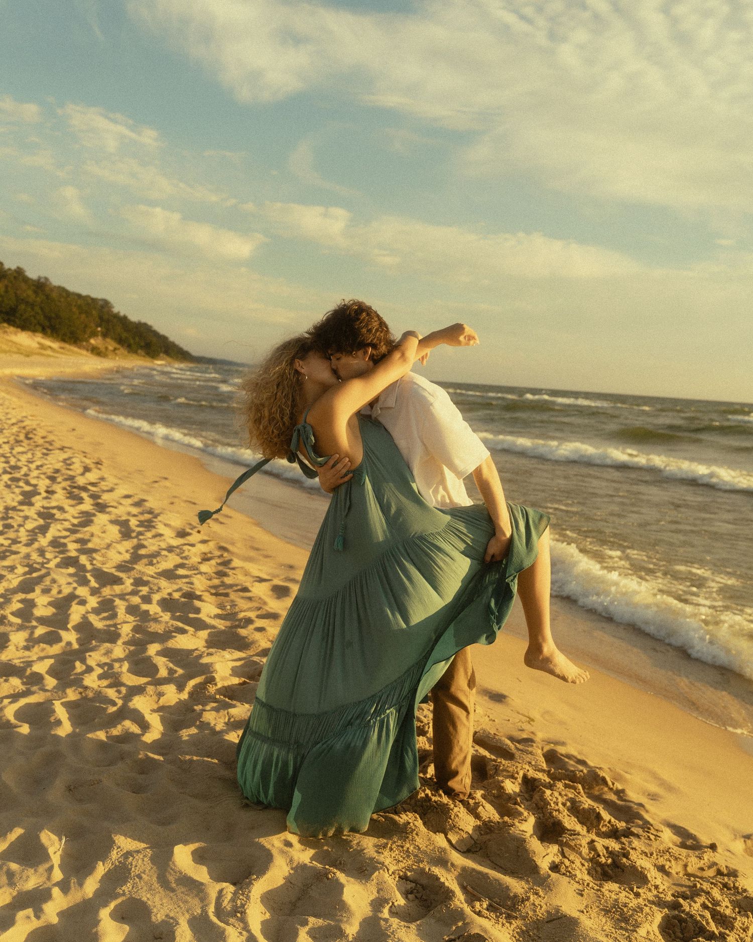 Couple sharing a romantic kiss on a sandy beach at sunset, woman in flowing green dress lifted off the ground.