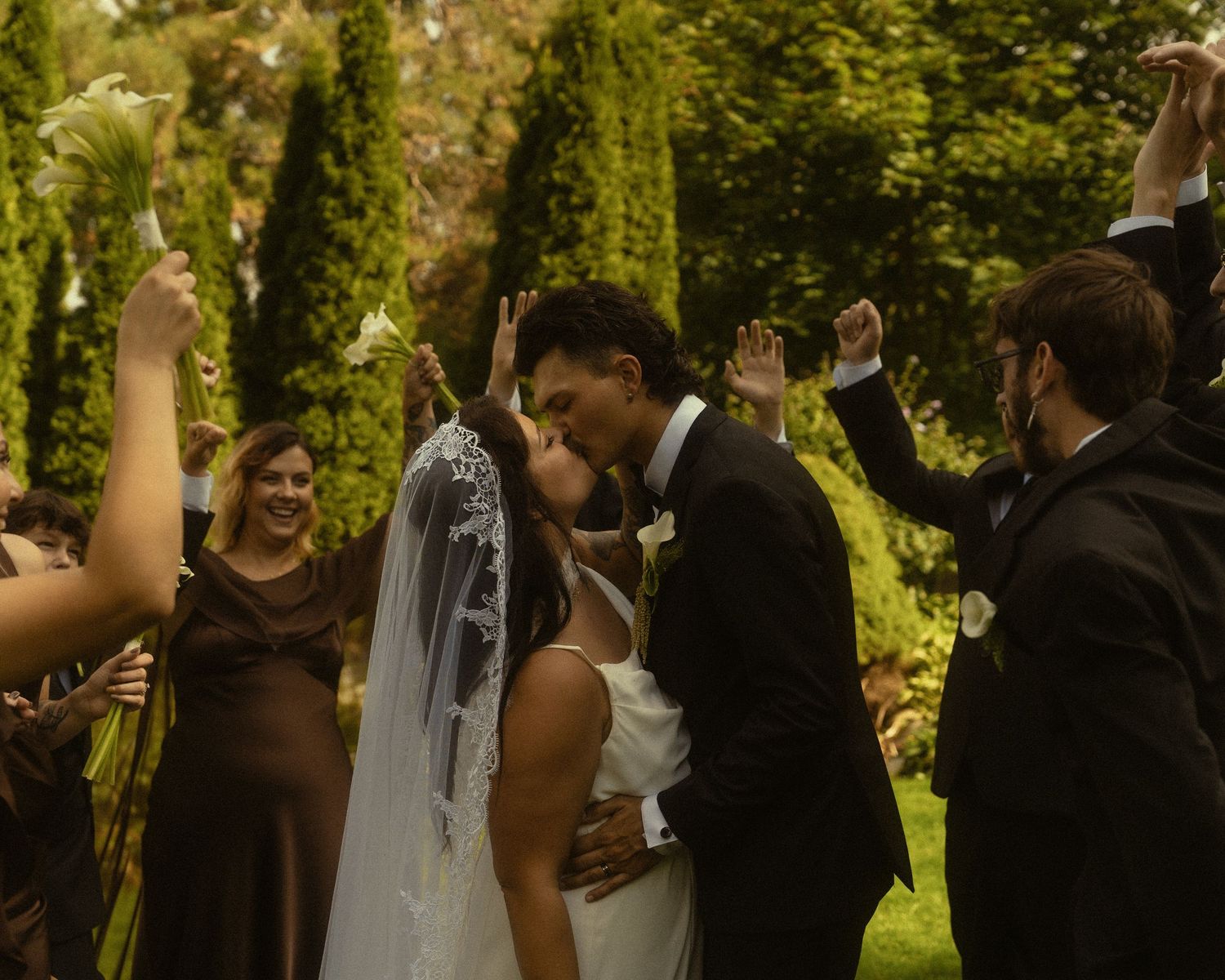 Bride and groom share first kiss at outdoor wedding ceremony surrounded by cheering guests in formal attire.