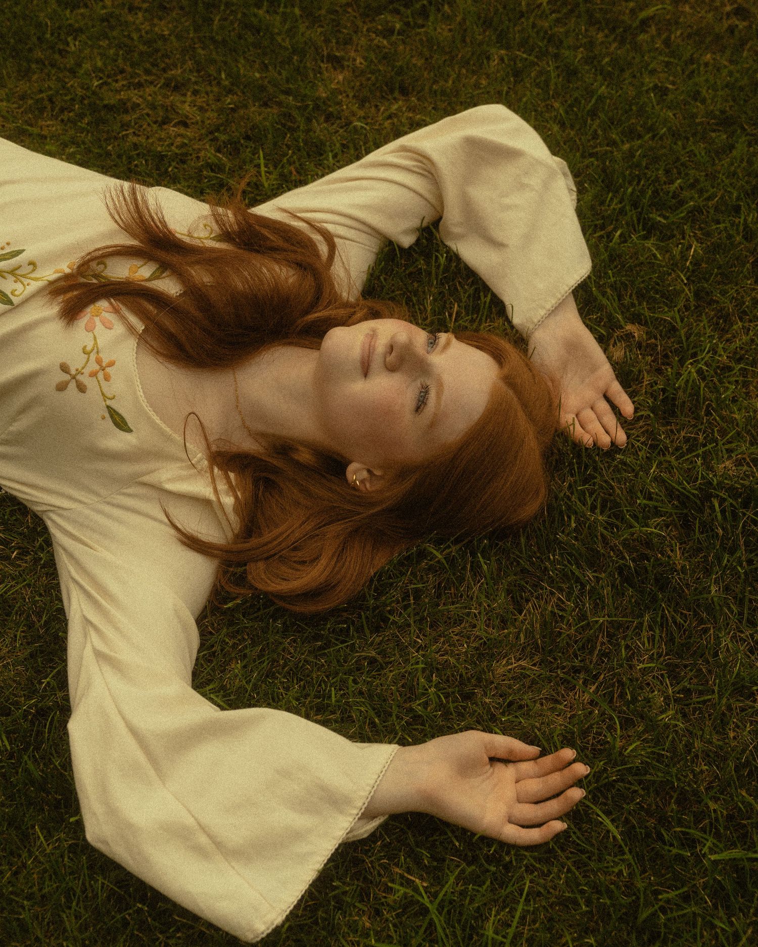 A red-haired woman in a white floral dress lies peacefully on green grass, eyes closed, arms outstretched.