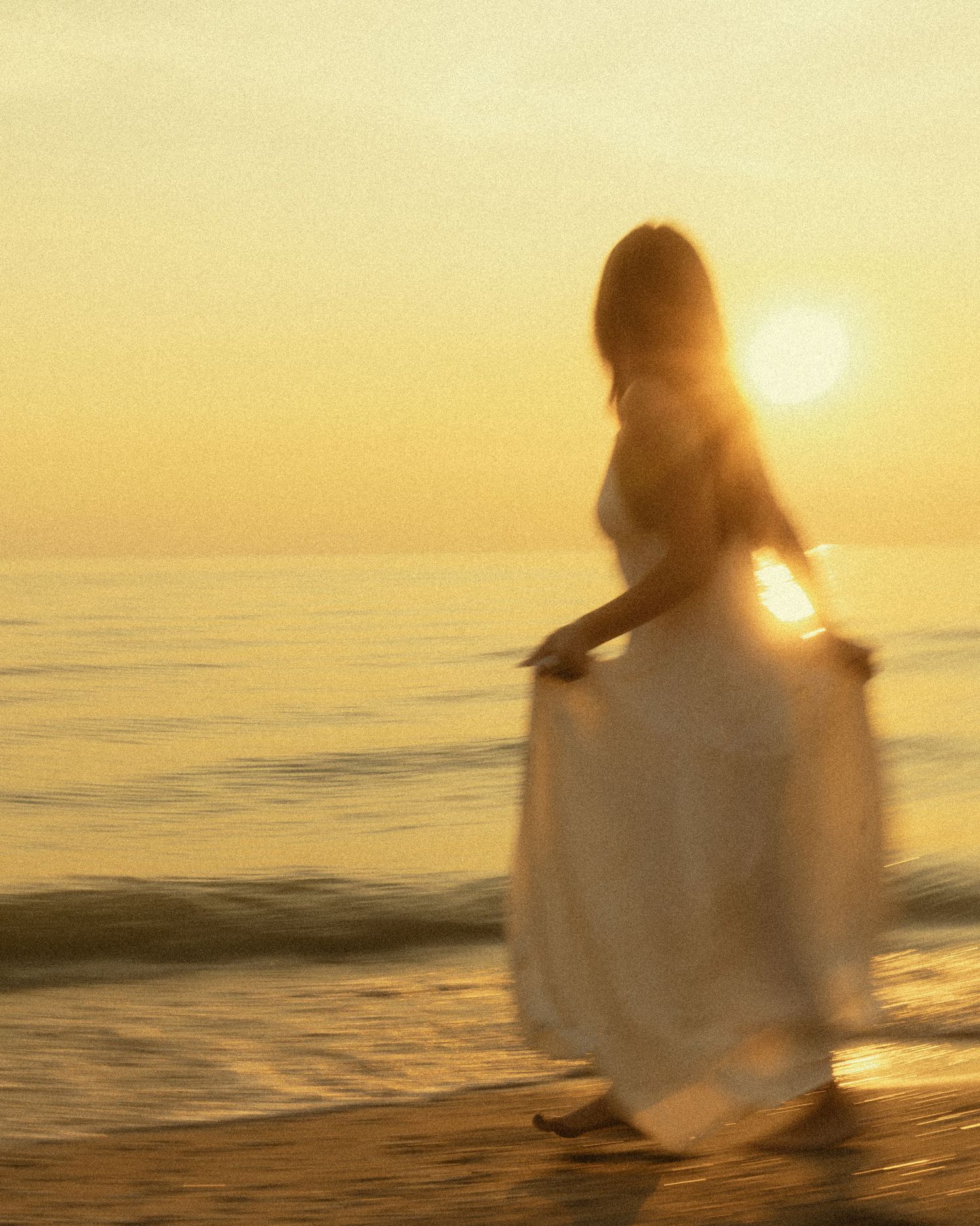 Woman in white dress walking on beach at golden sunset, waves lapping shore in warm glowing light.