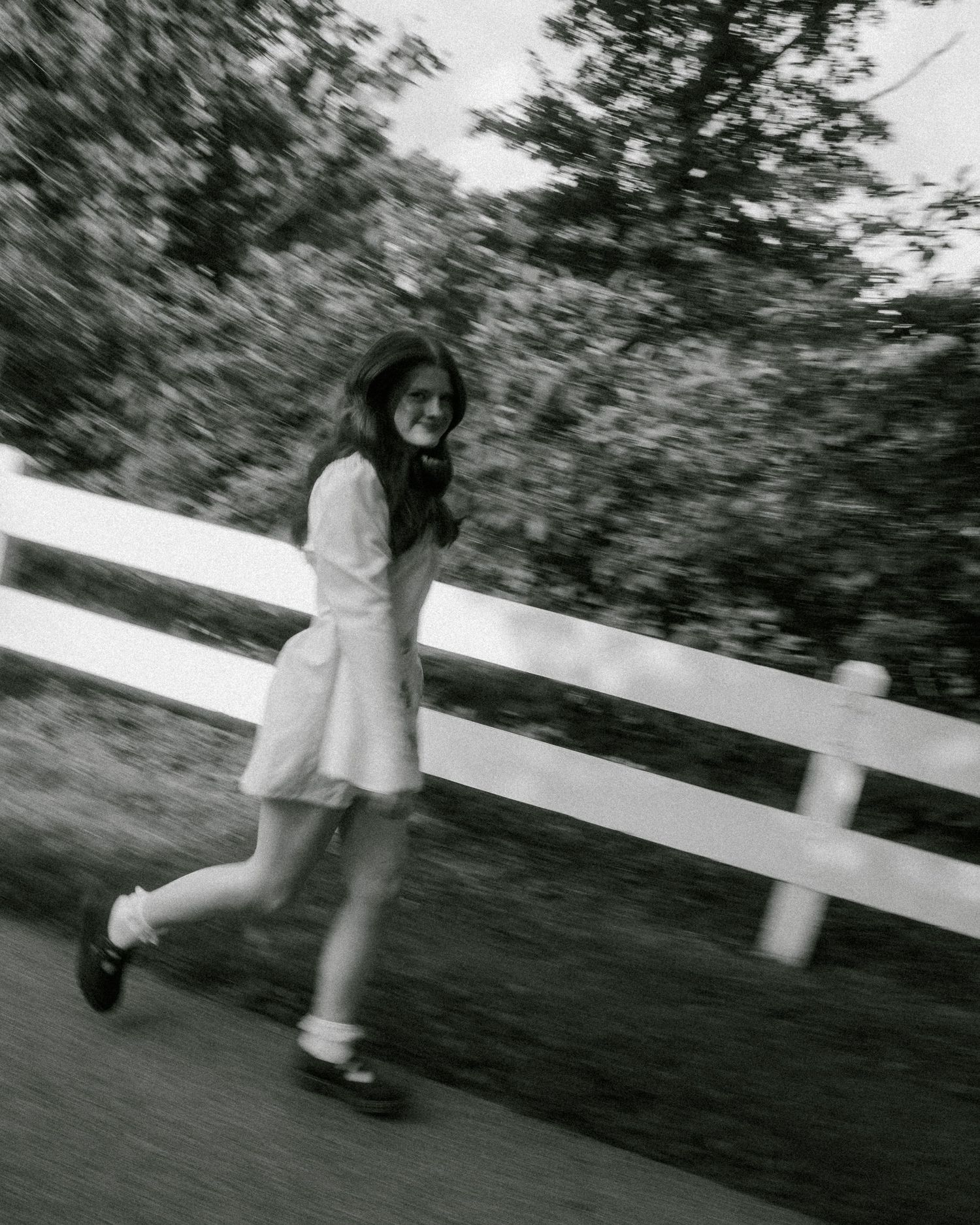 Black and white photo of a young woman in a white dress running along a path beside a white fence.
