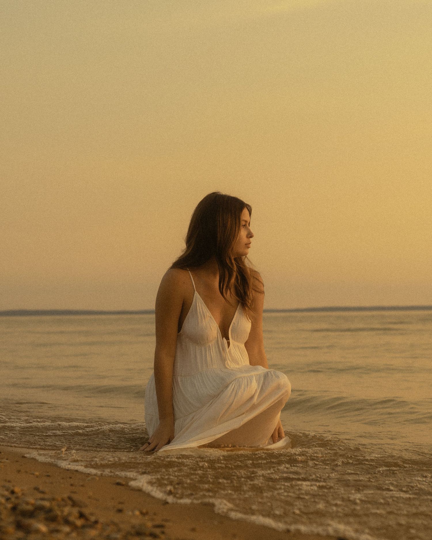 Woman in white dress sitting on sandy beach shore during golden sunset with calm ocean waves.