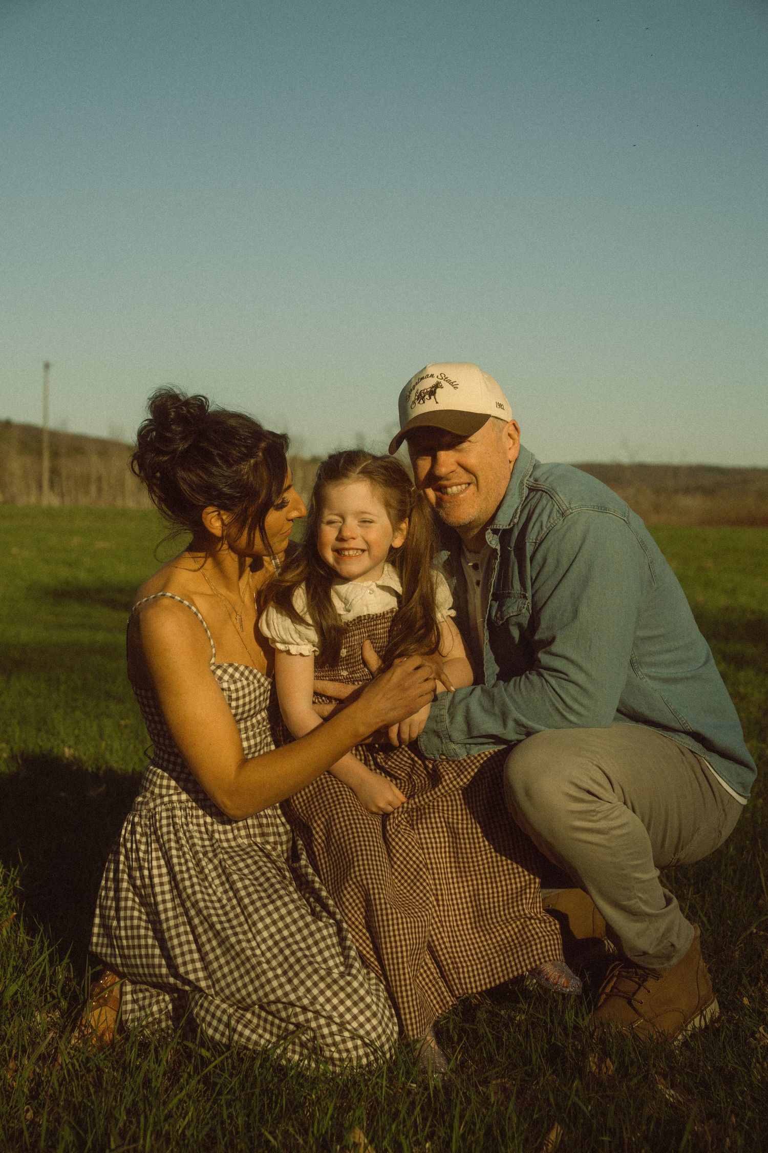 A happy family of three poses outdoors in golden hour light, with parents smiling at their young daughter in matching plaid outfits.