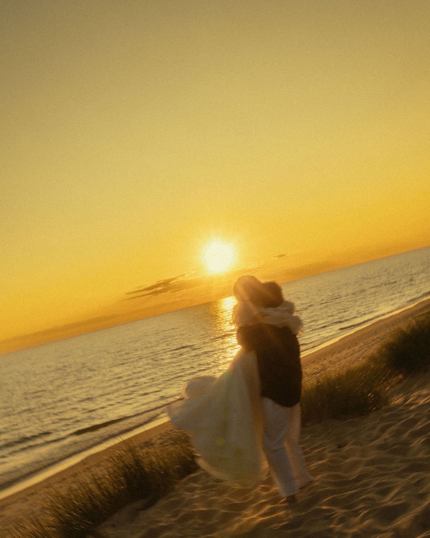 Couple embracing on a sandy beach at golden sunset, waves glistening, woman in flowing white dress.