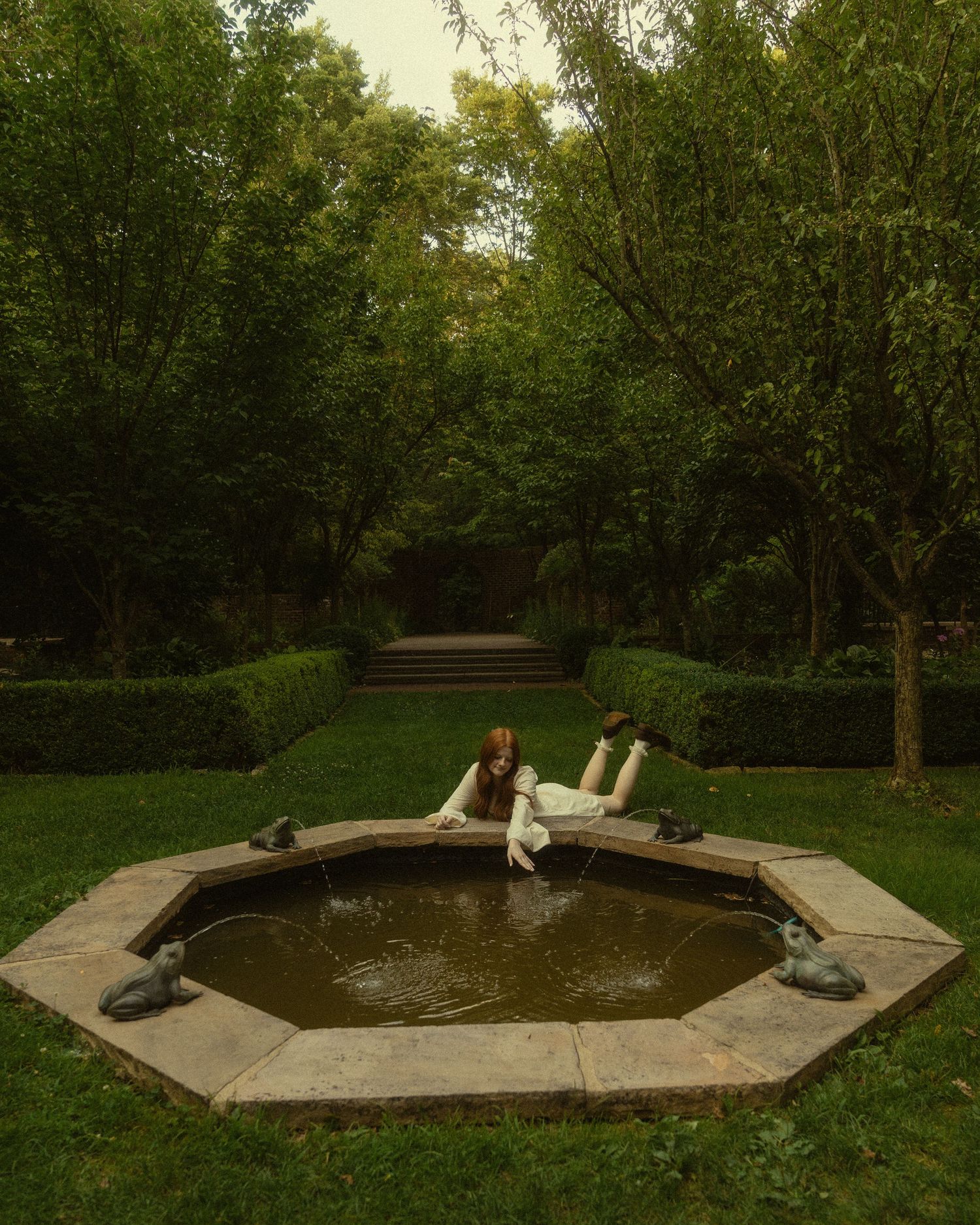 Two people relaxing by an octagonal garden fountain surrounded by lush green trees and manicured hedges.