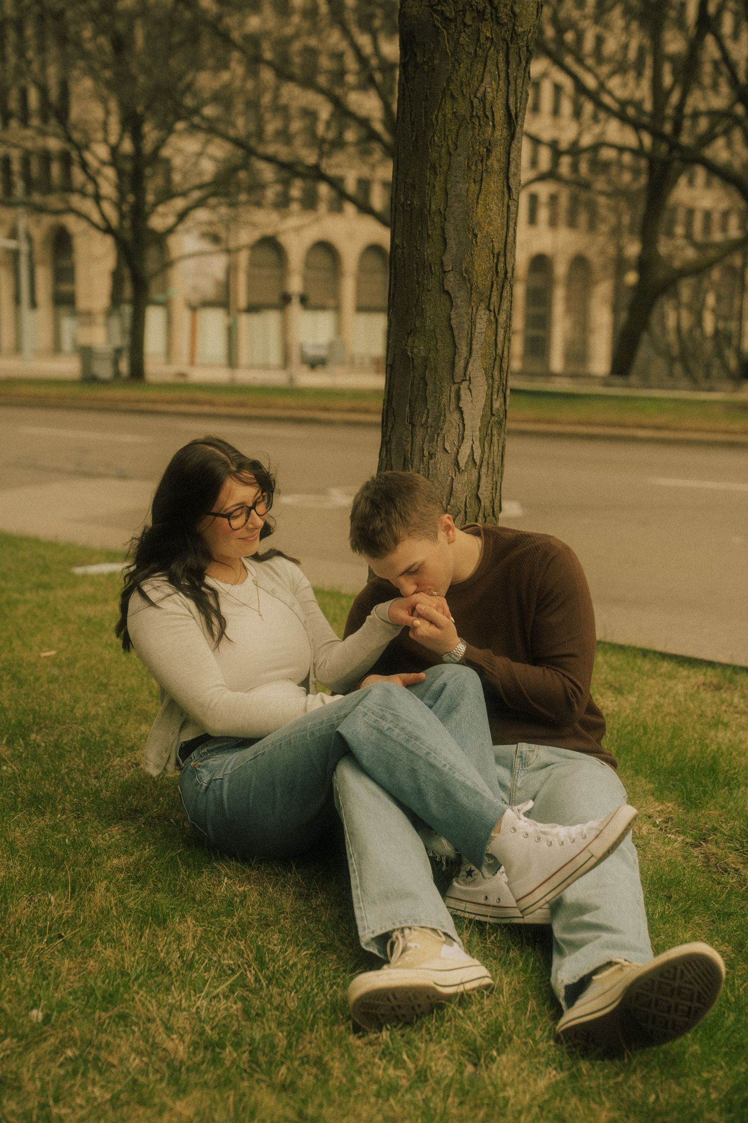 Couple sitting on grass by a tree, man kissing woman's hand, wearing jeans and casual tops outdoors.