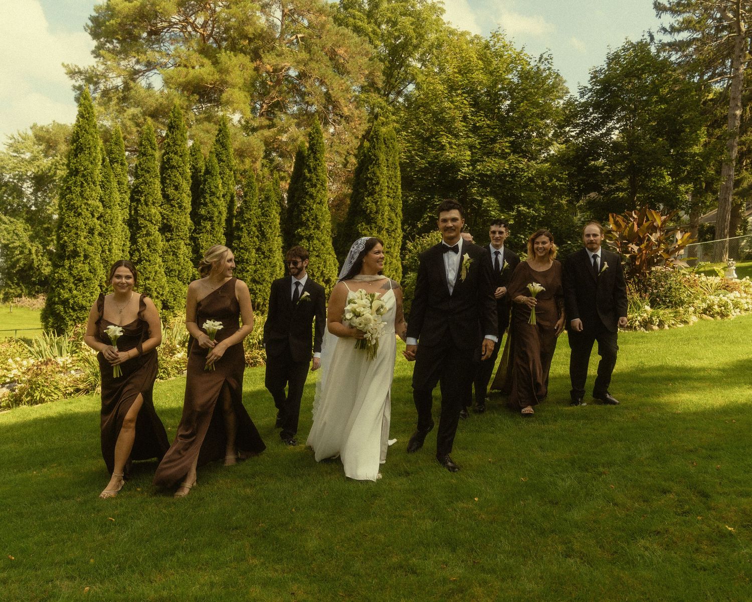 Wedding party walks on lush green lawn, bride in white gown, groom in tuxedo, bridesmaids in brown dresses, groomsmen in suits.