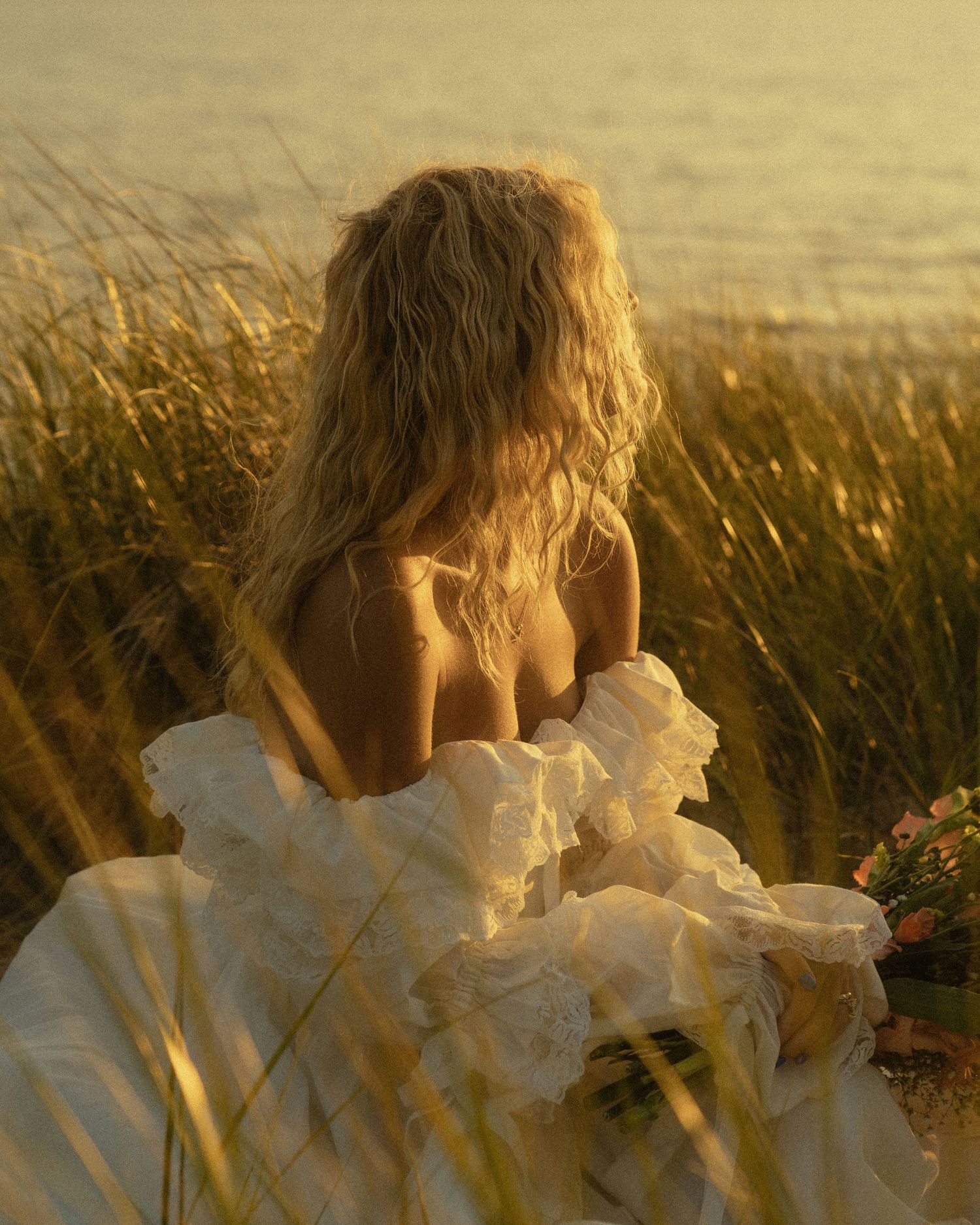 A blonde woman in a ruffled white gown holds flowers by golden beach grass at sunset.