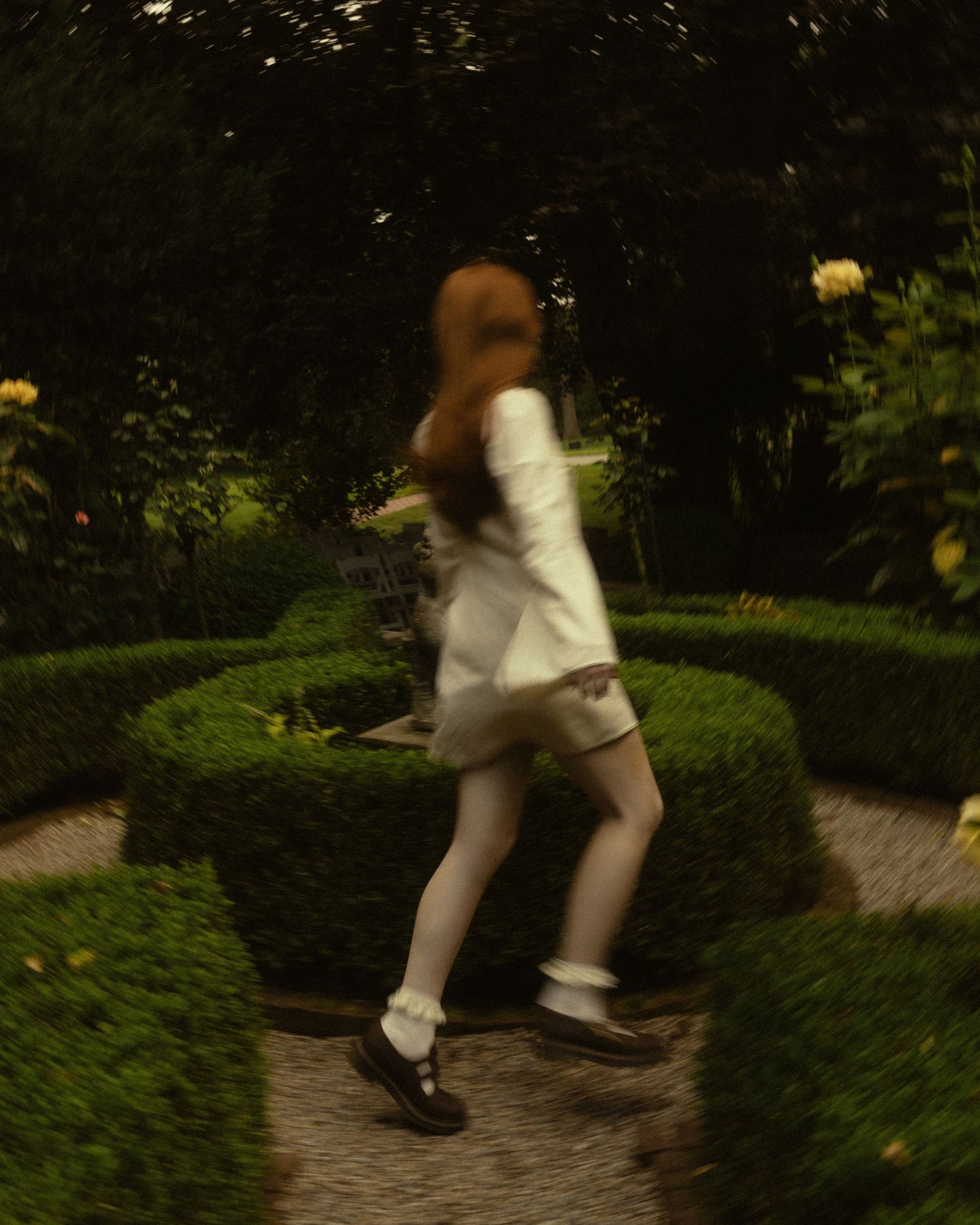 A blurred girl in white dress and mary janes runs through a lush green formal garden maze at dusk.