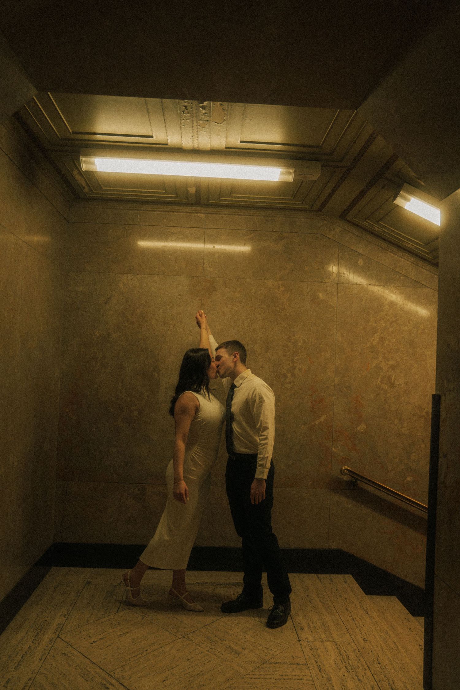 Couple sharing a romantic moment in an elegant elevator, woman reaching up while man leans in close.