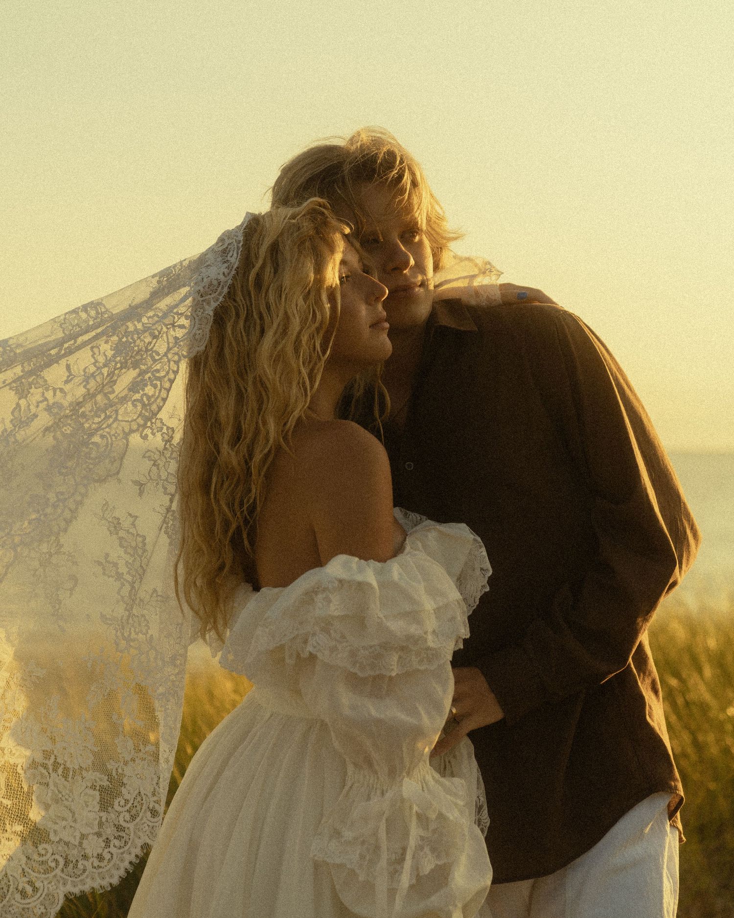 Couple sharing a romantic kiss at golden hour, bride in white lace dress with veil flowing in the breeze.