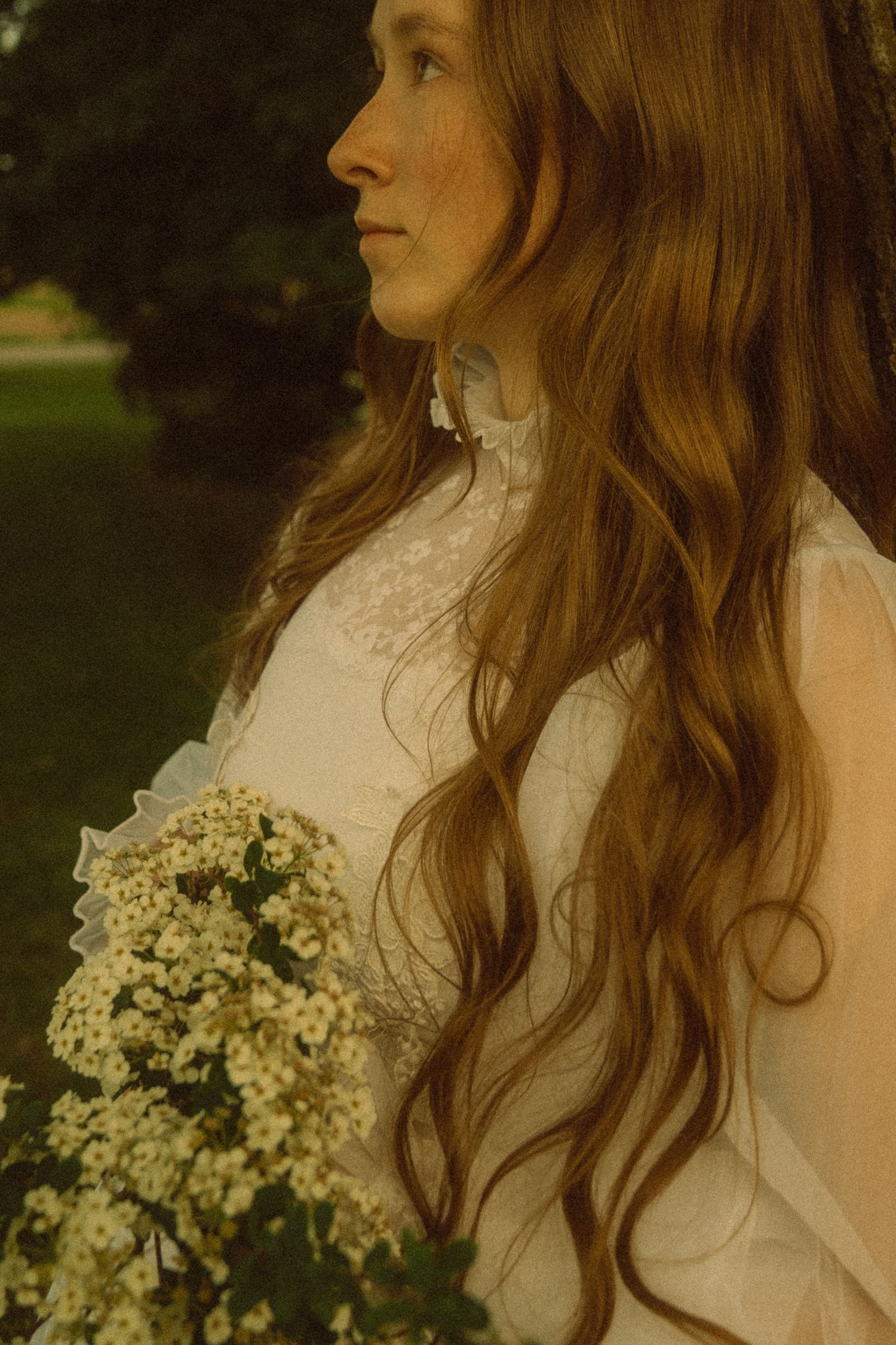A young woman with long wavy auburn hair in a white lace dress holds a bouquet of white wildflowers outdoors.