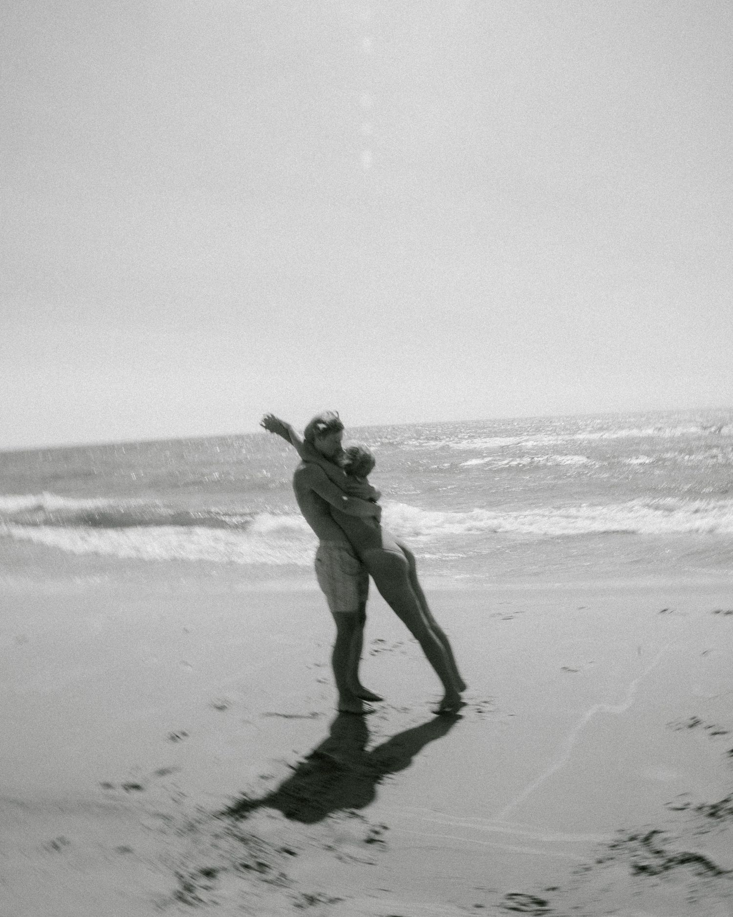 Black and white photo of a couple embracing on a wet beach with ocean waves in the background.