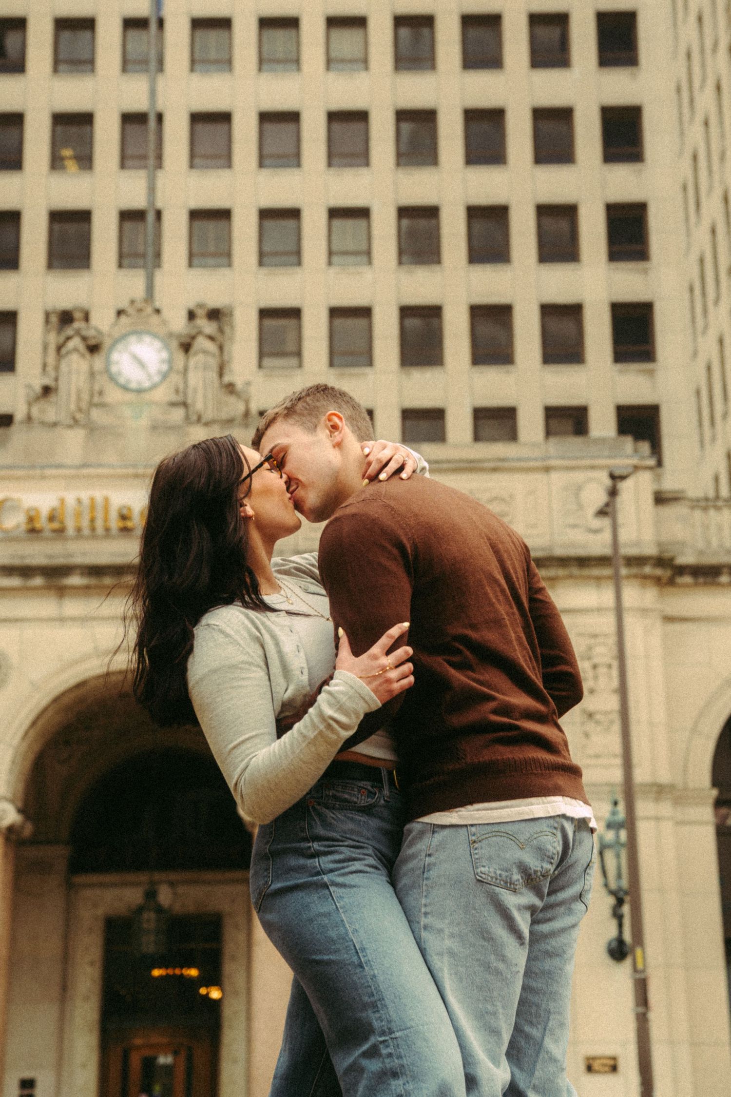 A couple shares a romantic kiss in front of an urban building with classical architecture.