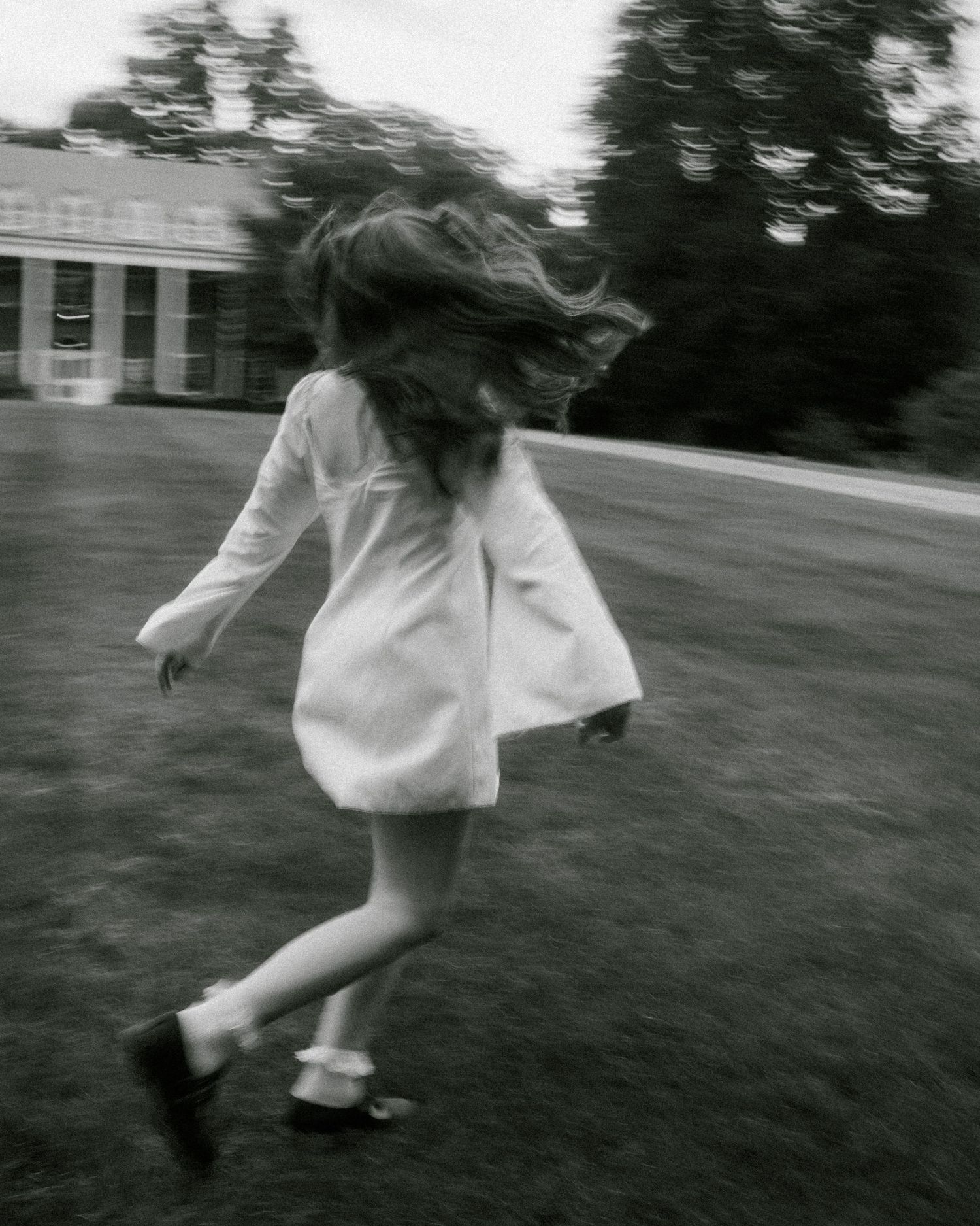 Black and white photo of a girl in a white dress running across a lawn, her long hair flying behind her.