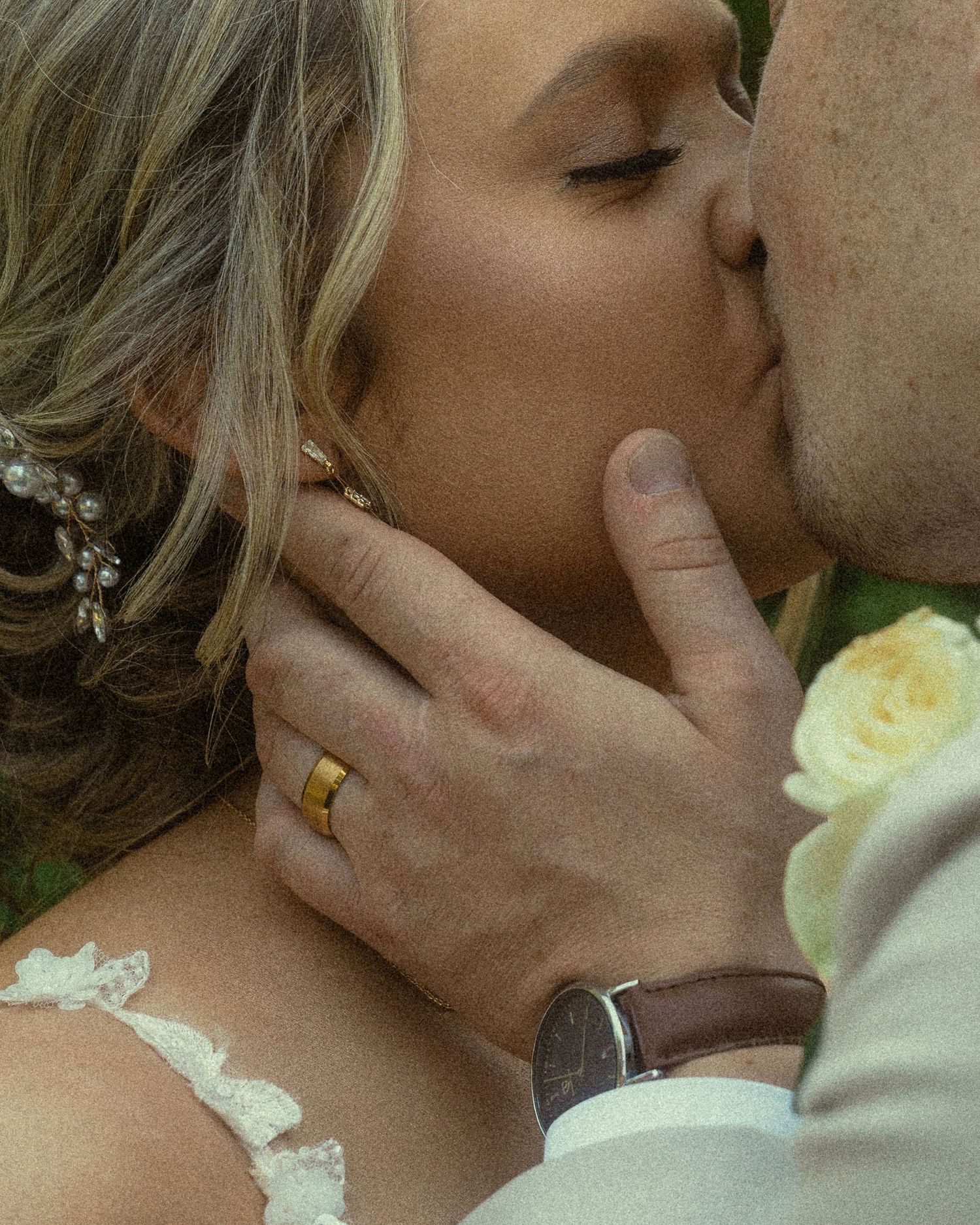 Close-up of a bride and groom sharing a kiss, wedding rings visible, with white roses in the background.