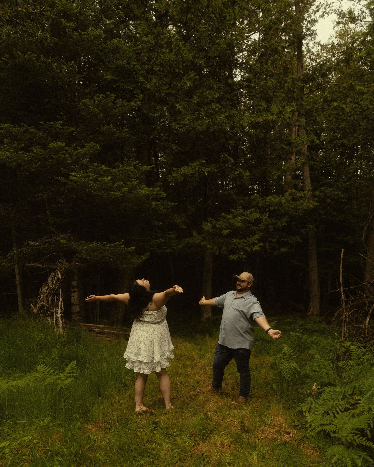 Couple dancing in a lush green forest clearing, woman in white dress with arms outstretched, man in gray shirt smiling.