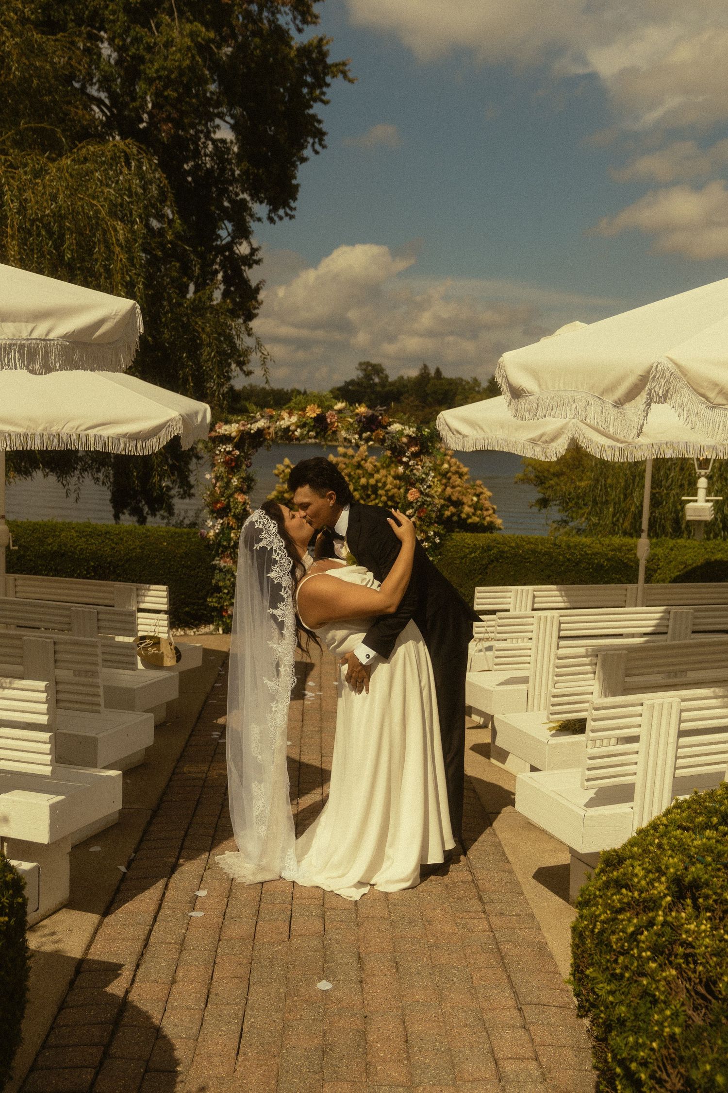 Bride and groom share first kiss at outdoor wedding ceremony with white chairs and umbrellas near a lake.