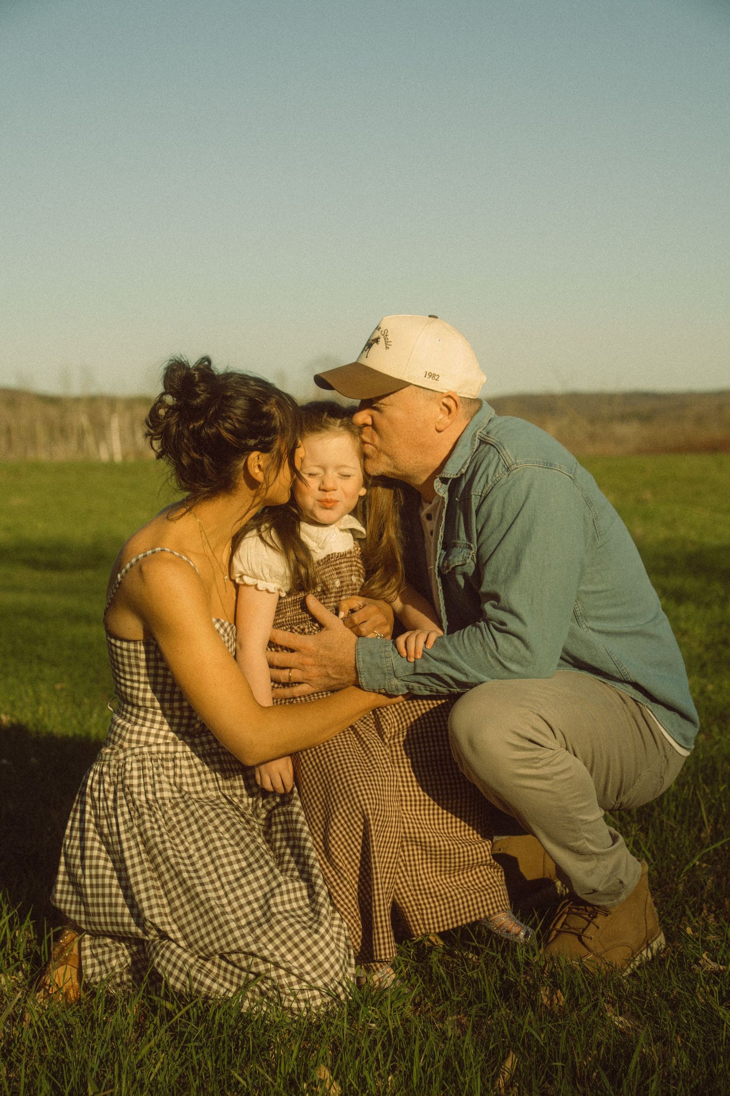 A family of three shares a tender moment in a sunny green field, with parents kissing their toddler.