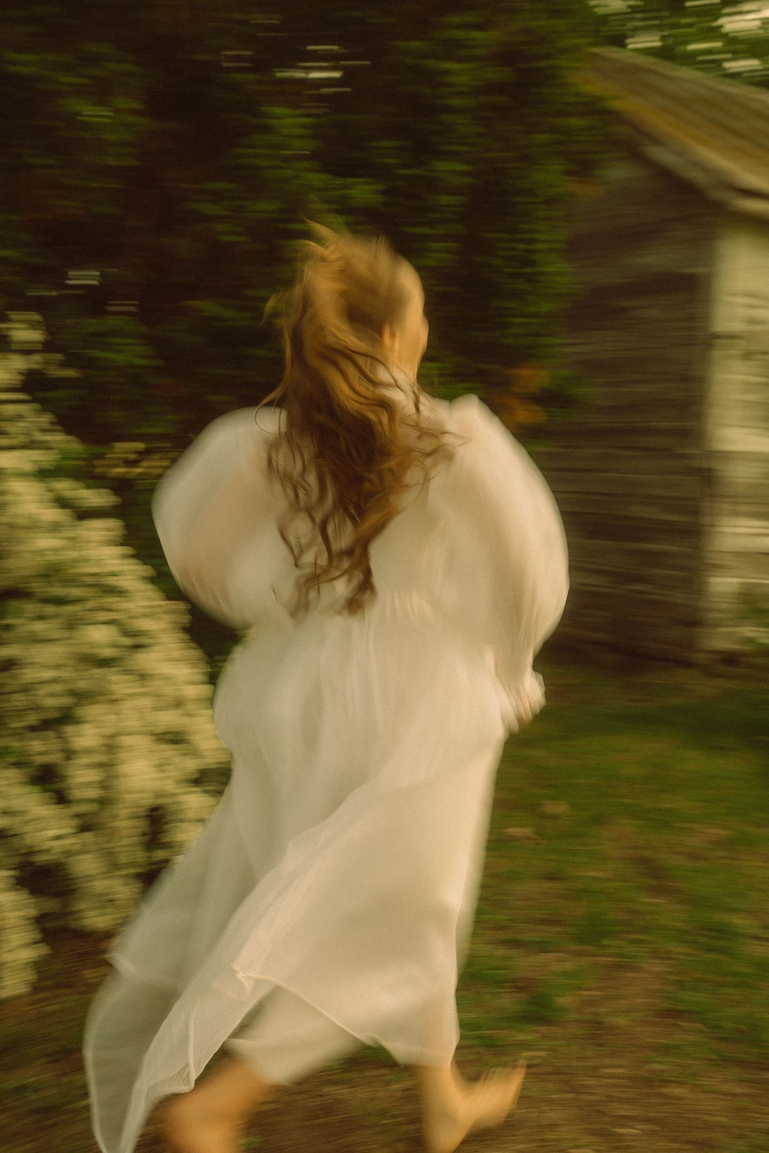 A woman in a flowing white dress runs barefoot through a lush garden toward a rustic wooden shed.