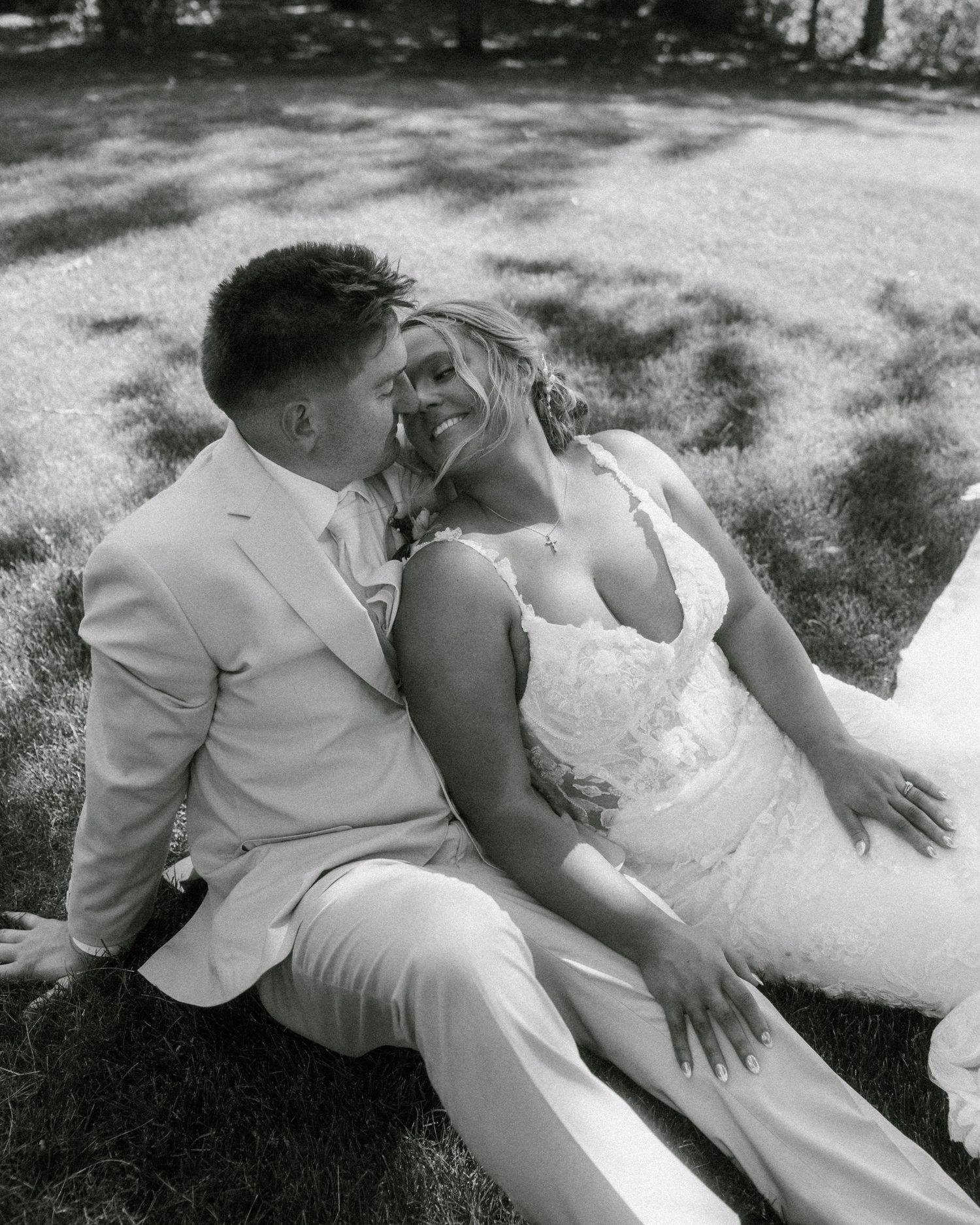 Black and white photo of a bride and groom sitting on grass, smiling at each other on their wedding day.