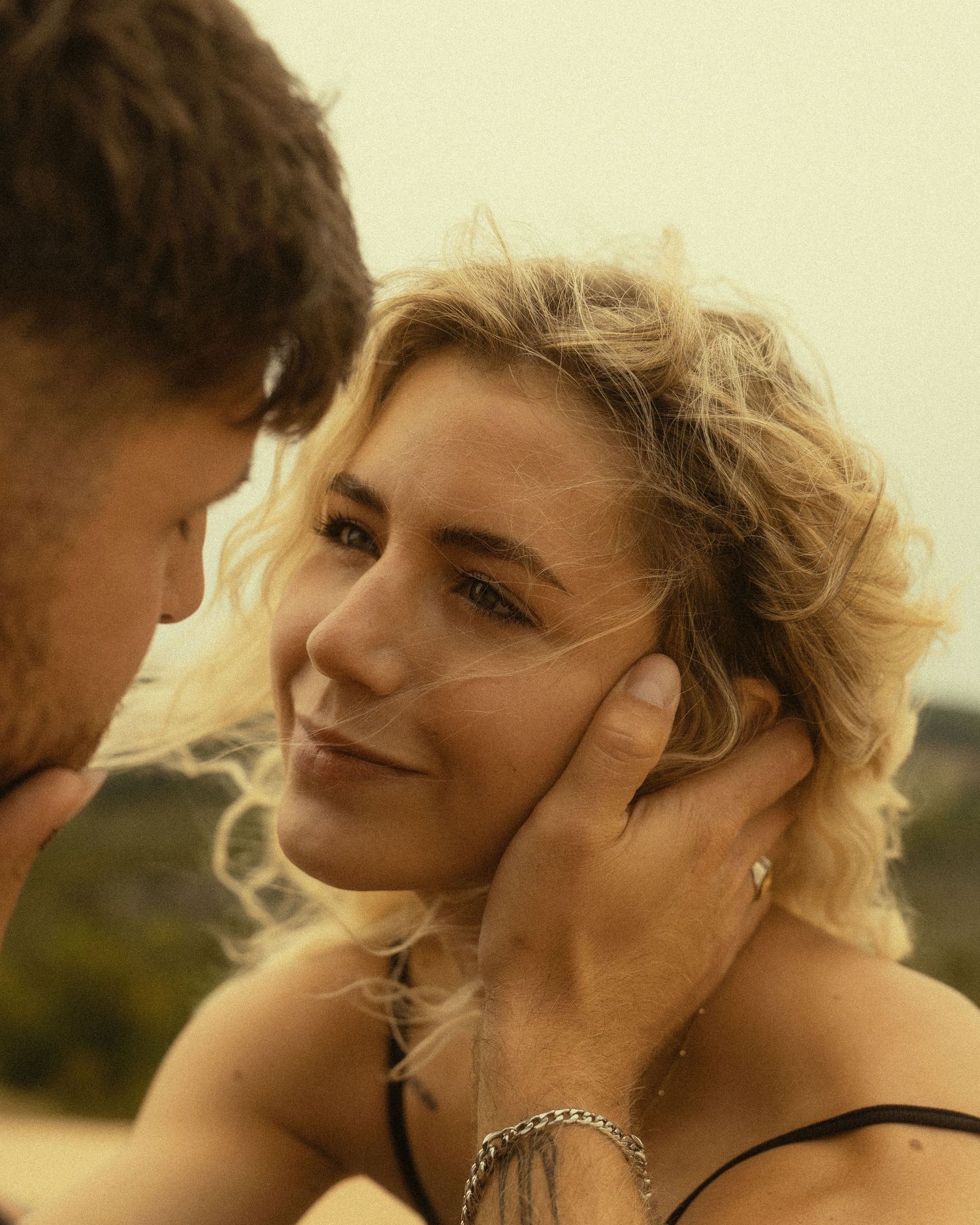 A blonde woman smiles warmly as a man gently cups her face, sharing an intimate romantic moment outdoors.