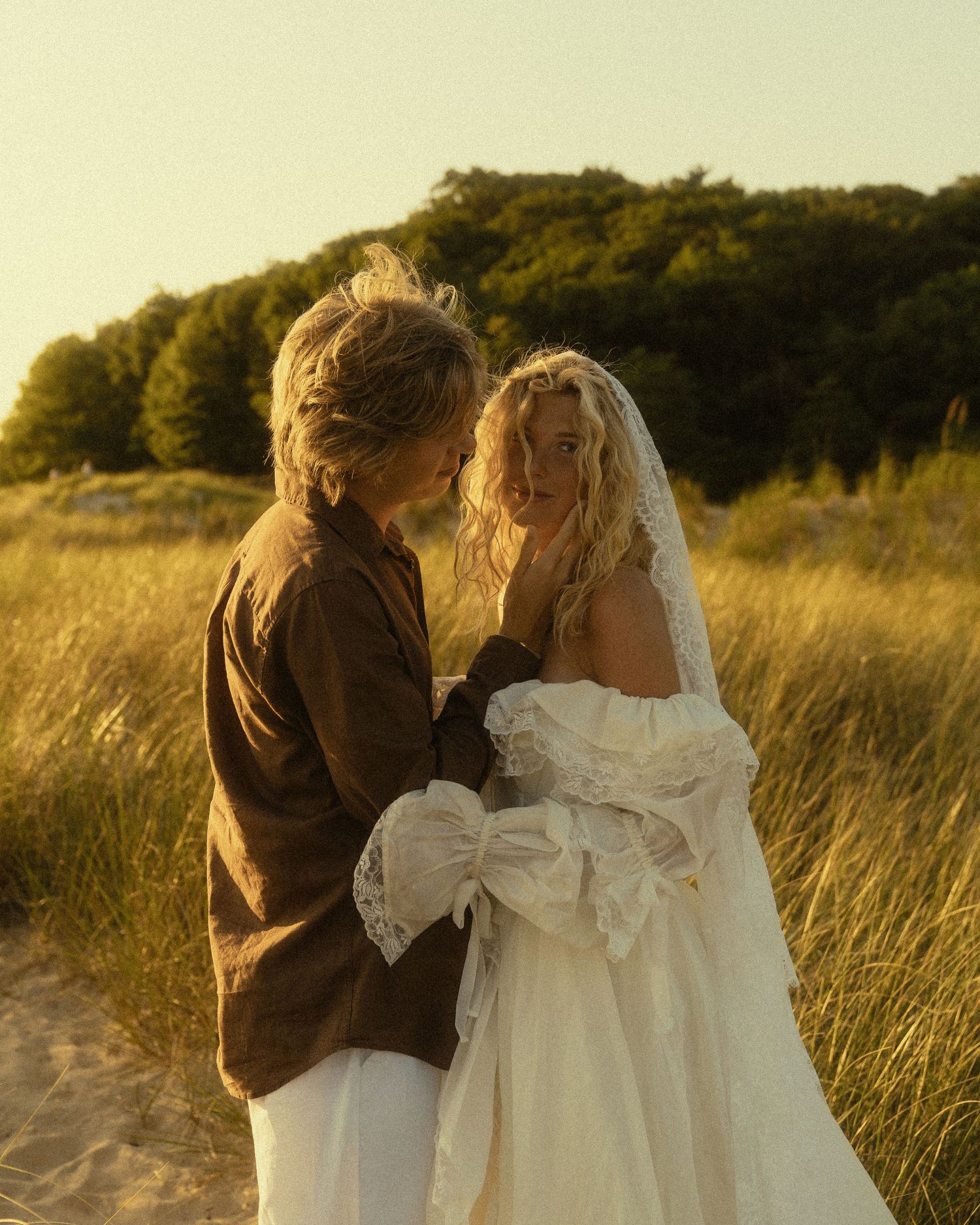 Couple in wedding attire embracing on sandy dunes at golden hour with lush green trees in background.