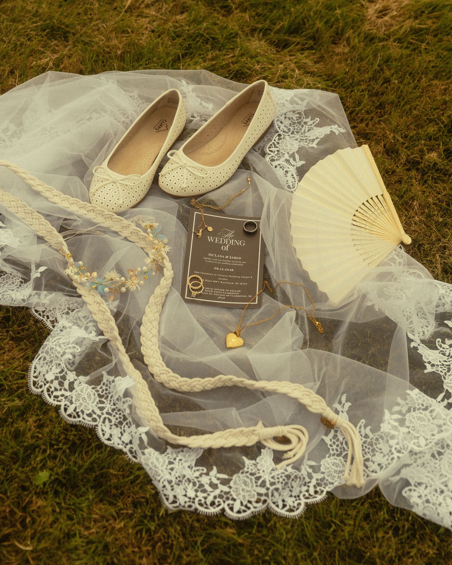 Wedding flat shoes, lace veil, hand fan, invitation, pearl necklace, and gold rings arranged on green grass.