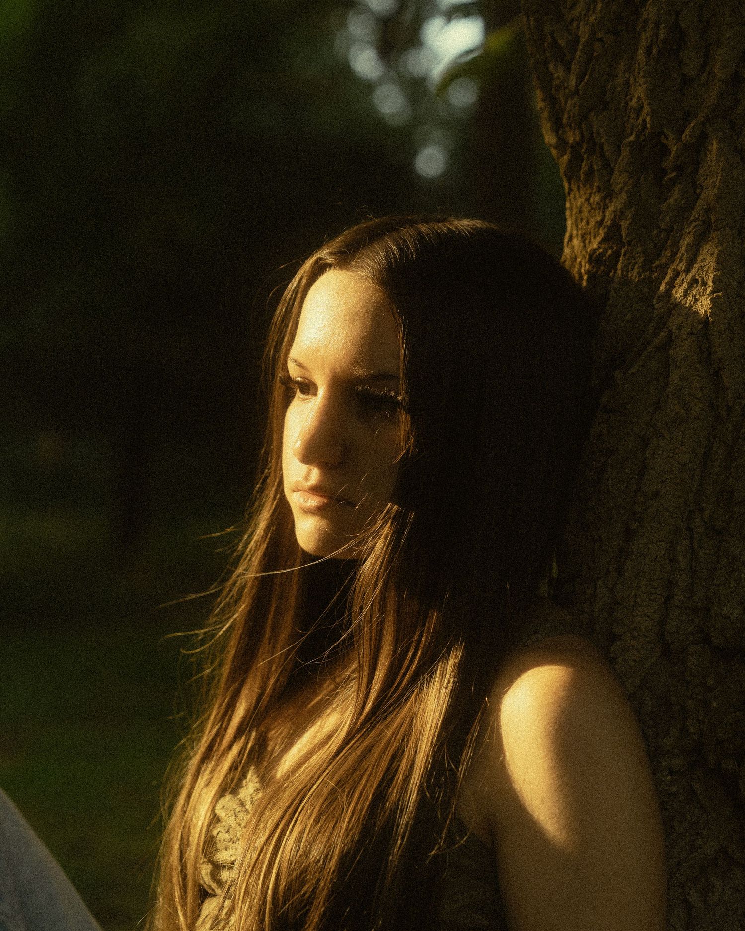 Young woman with long brown hair leaning against a tree in golden hour sunlight in a lush green forest.