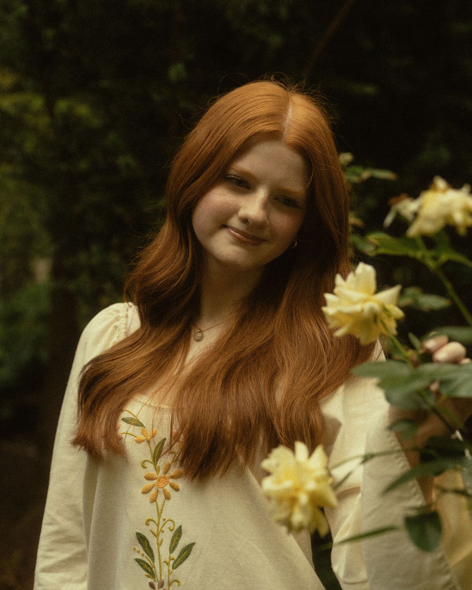 Young woman with long red hair smiles among yellow roses, wearing a white floral embroidered top outdoors.