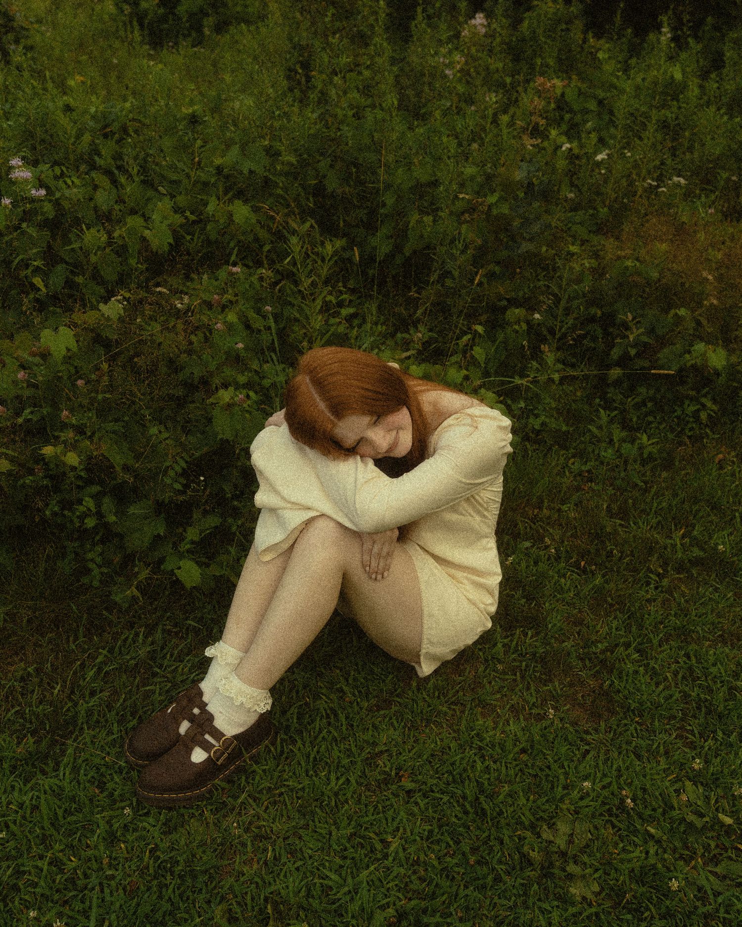 A redhead woman in cream outfit sits curled up on green grass, hugging her knees, wearing brown shoes.