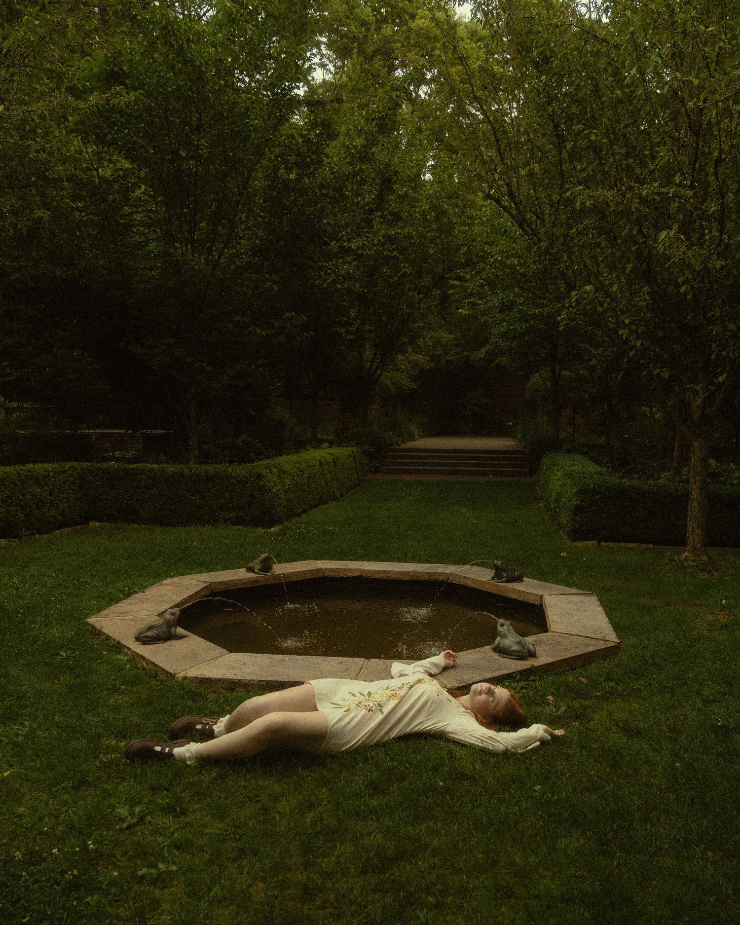 A person in white dress lying on grass beside an octagonal garden pond surrounded by lush green trees.